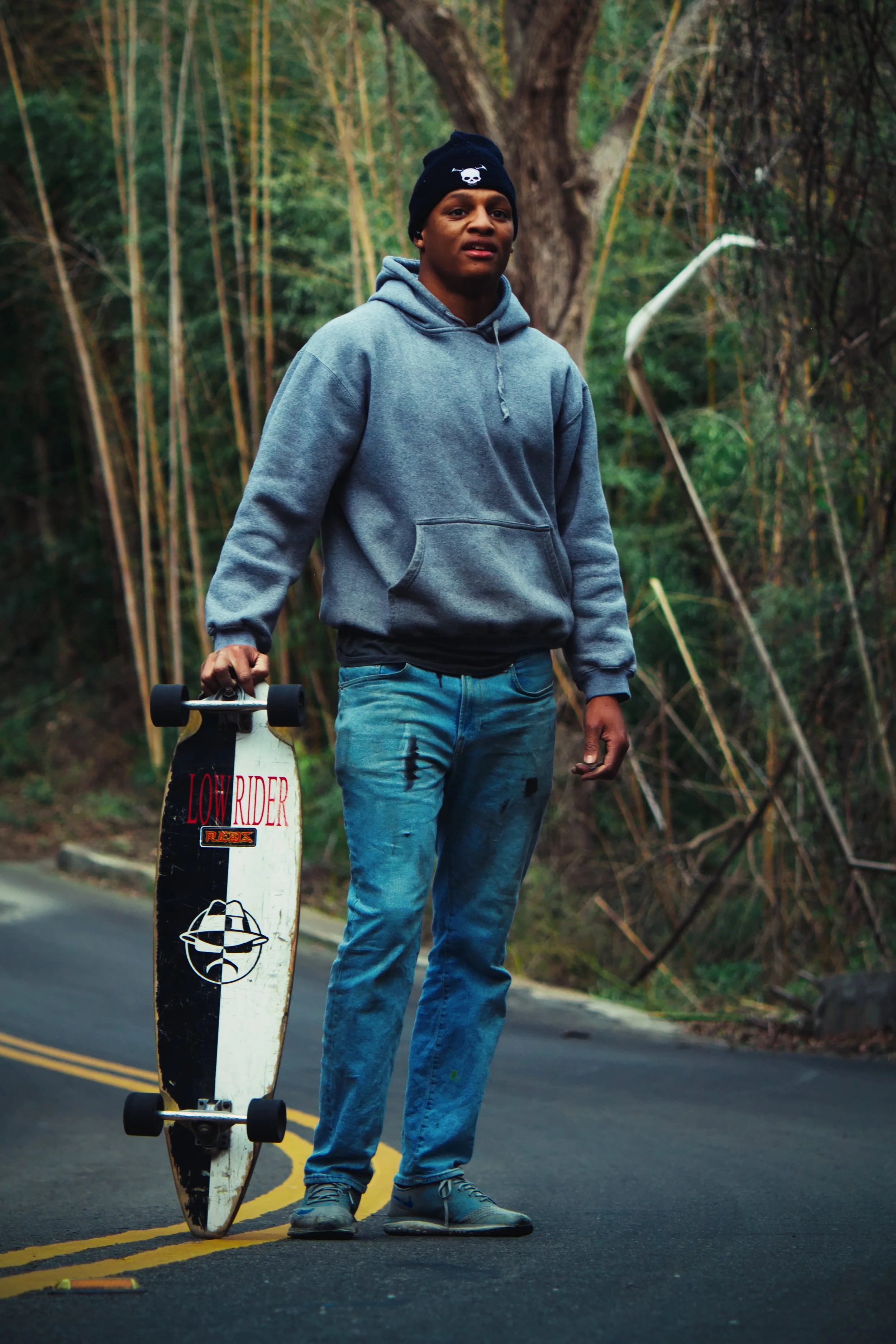 Young man wearing a gray hoodie and jeans, holding a skateboard, standing on a two-lane road surrounded by trees.