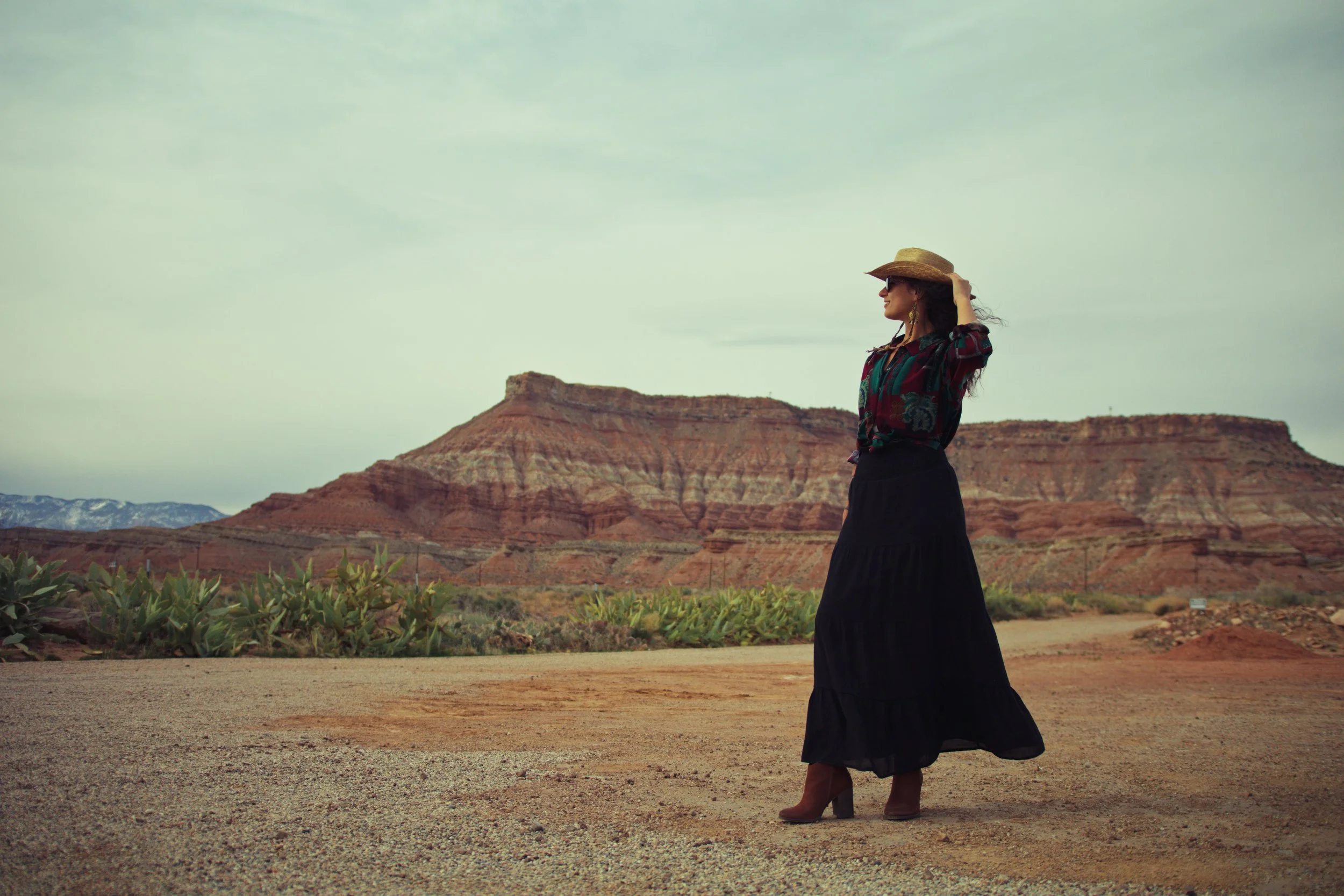 A woman standing on a dirt path in a desert landscape with red rock formations, wearing a wide-brimmed hat, sunglasses, a colorful blouse, a long black skirt, and boots.