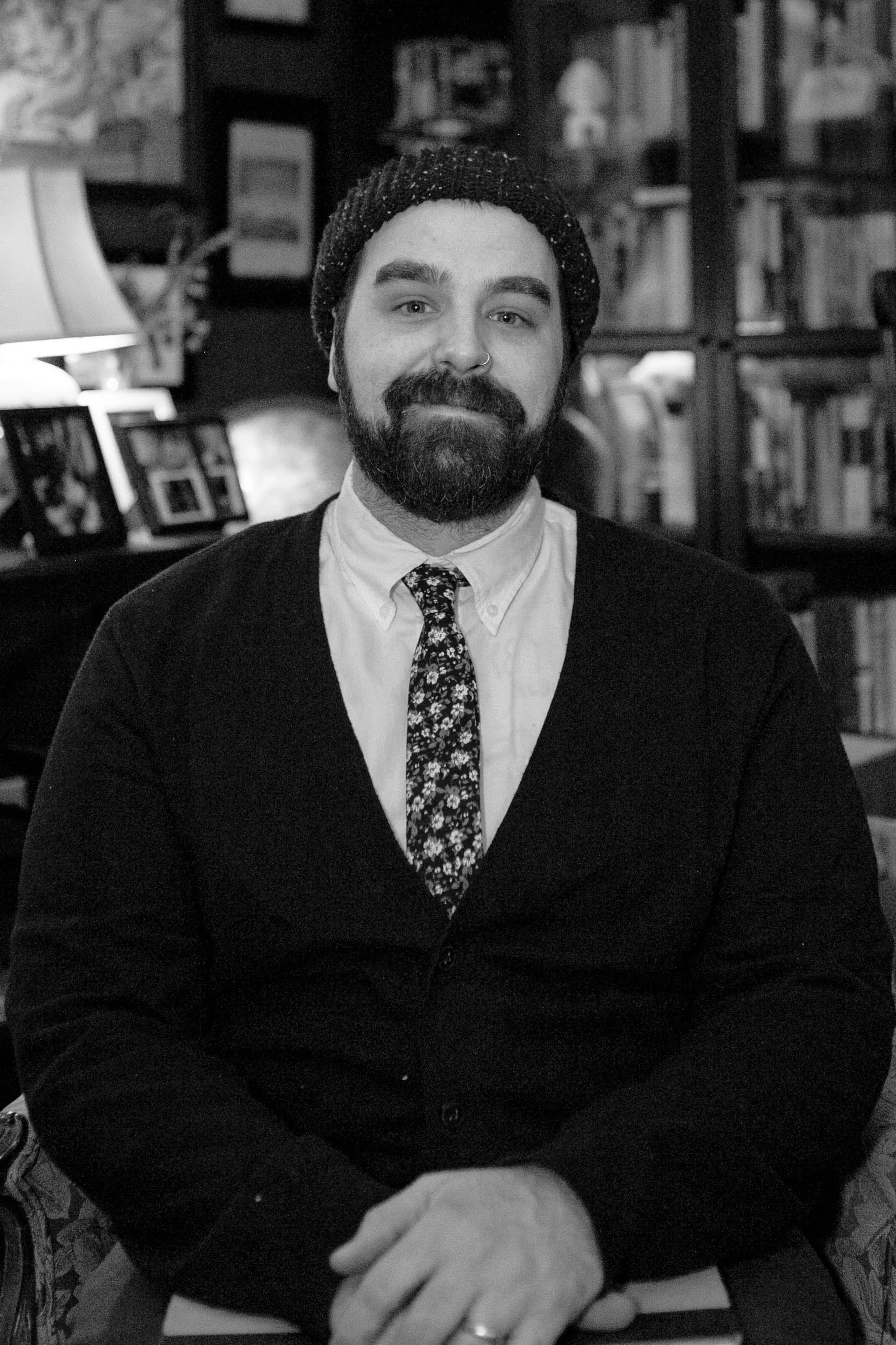 The poet Sean Early, known artistically as Uncle Zoid, smiles for the camera in a black and white portrait. He sports a beard and a nose ring and is dressed in a collared shirt, cardigan, tie and a beanie.