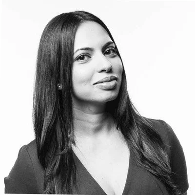 A woman with long dark hair, wearing a dark top, posing for a portrait against a plain light background.