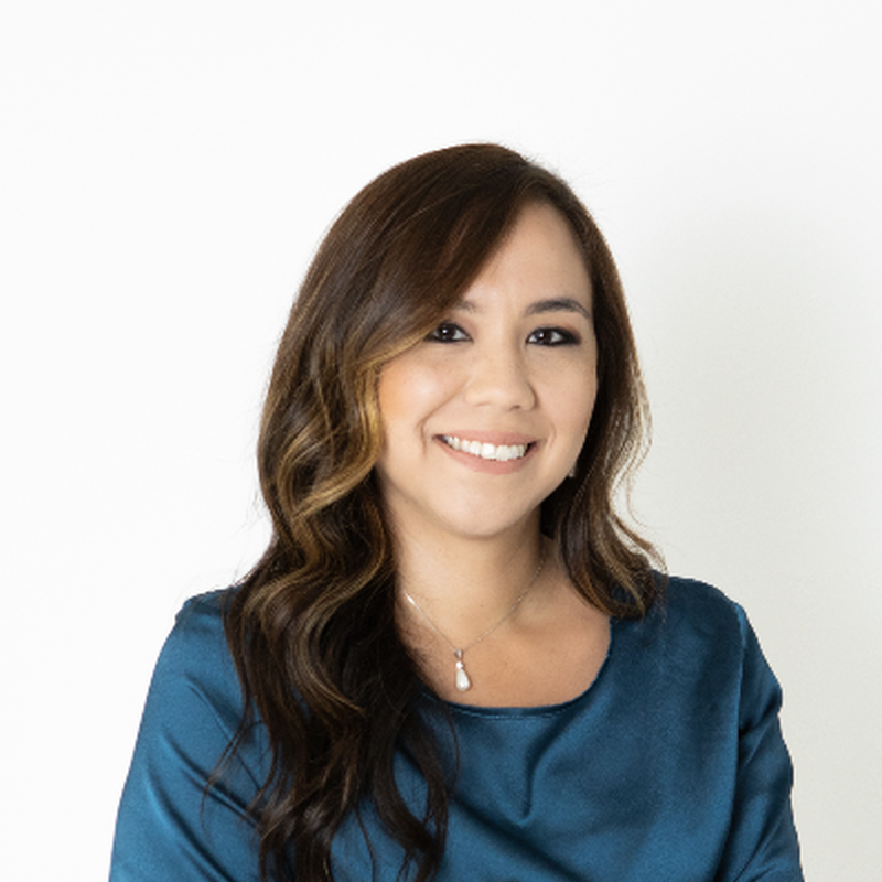 A woman smiling with long wavy brown hair wearing a blue top and a pendant necklace against a white background.