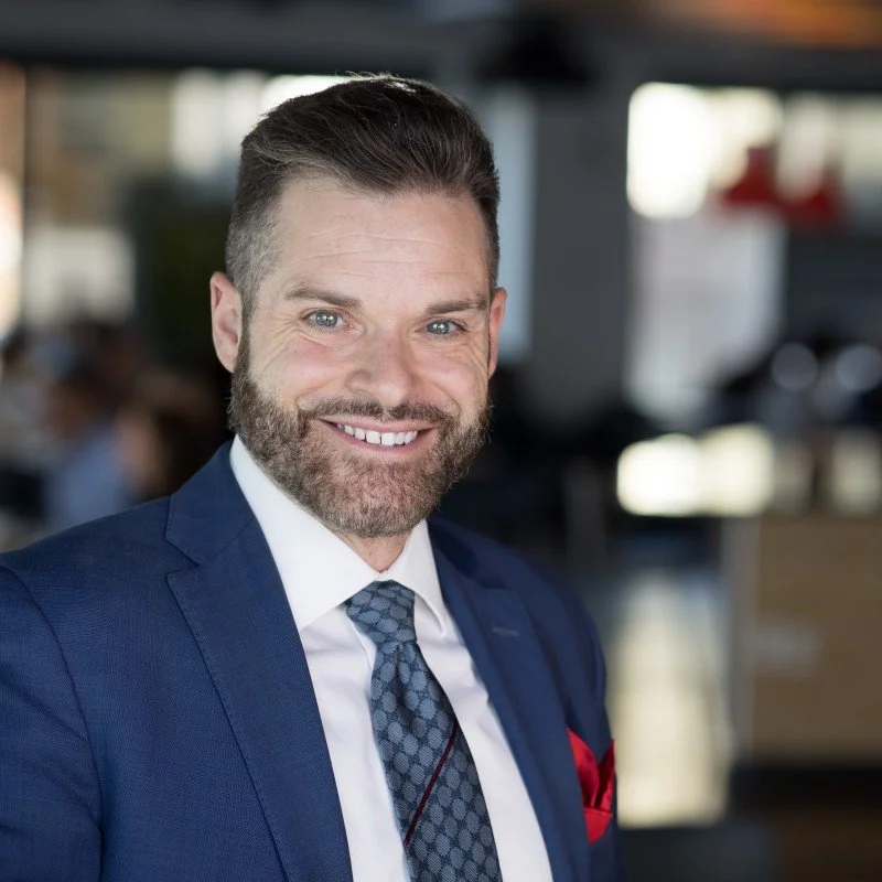 A smiling man with a beard and short hair, wearing a navy suit, white shirt, and patterned tie, in an indoor setting.