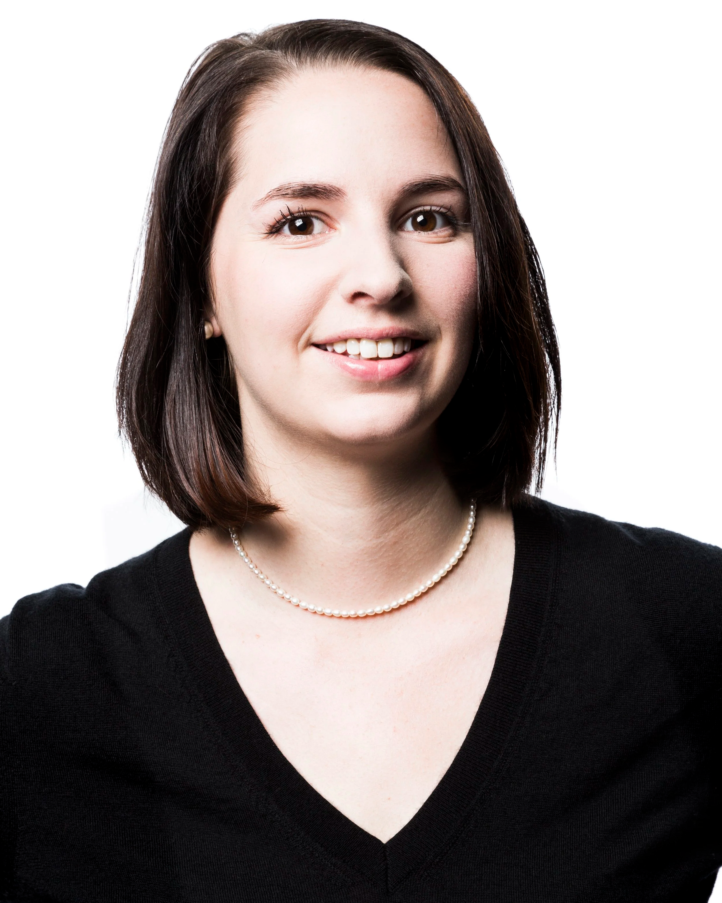 Close-up portrait of a young woman with shoulder-length dark hair, wearing a black V-neck top and a pearl necklace, smiling against a white background.