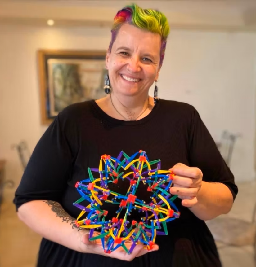 A smiling woman with short, colorful rainbow hair holding a complex, colorful geometric sculpture made of interconnected pieces.