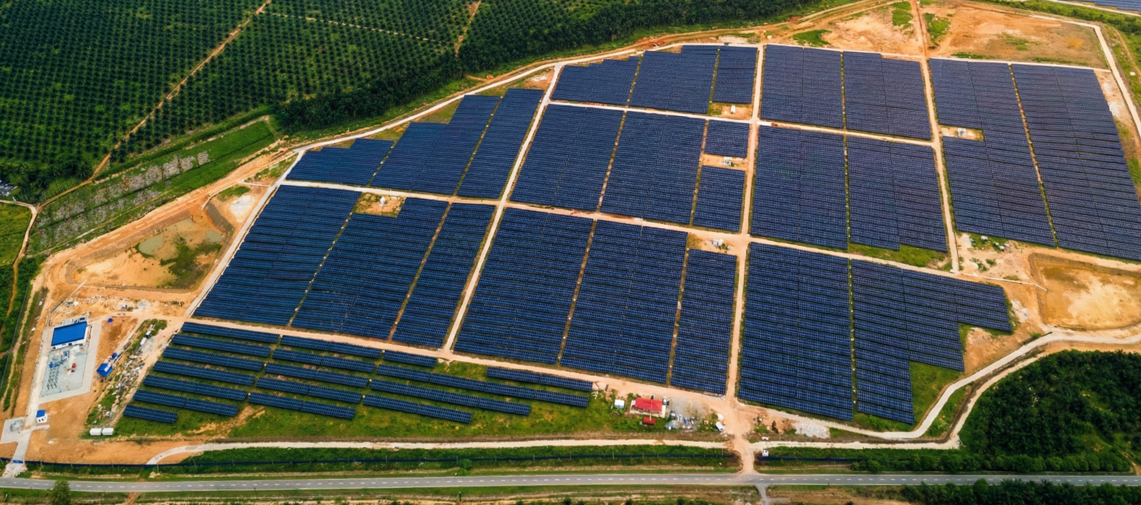Aerial view of a large solar panel farm with multiple rows of blue solar panels arranged in a grid, surrounded by dirt roads and patches of greenery, with a road at the bottom of the image.