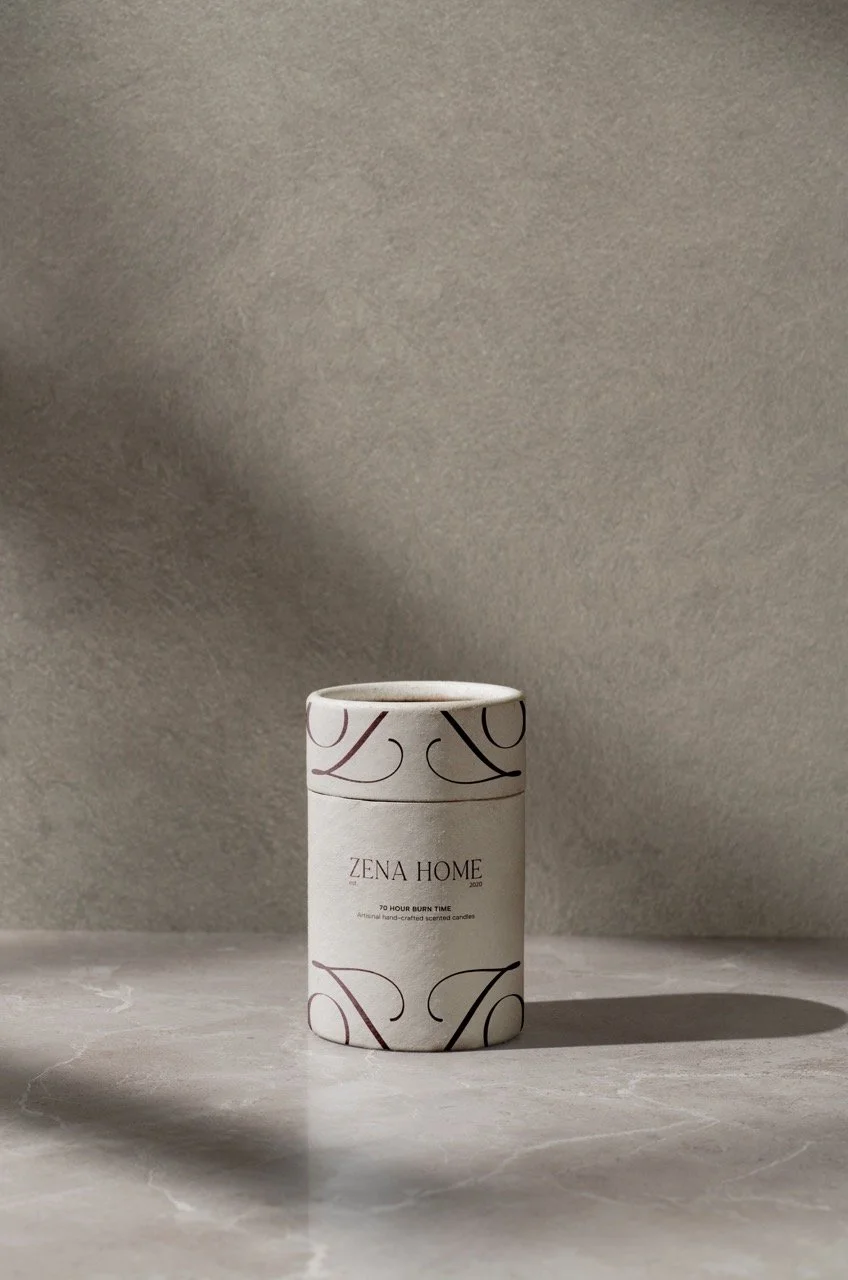 A white cylindrical candle container with artistic black and gold swirling lines, labeled 'Zena Home' and '70 Hour Burn Time,' placed on a marble surface with a plain neutral wall background.