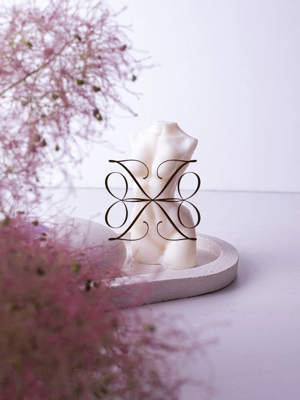 Decorative sculpture of a human torso on a white round base with a black, intricate, symmetrical metal art piece in front of it. Pink flowers are blurred in the foreground on the left, with a plain white background.