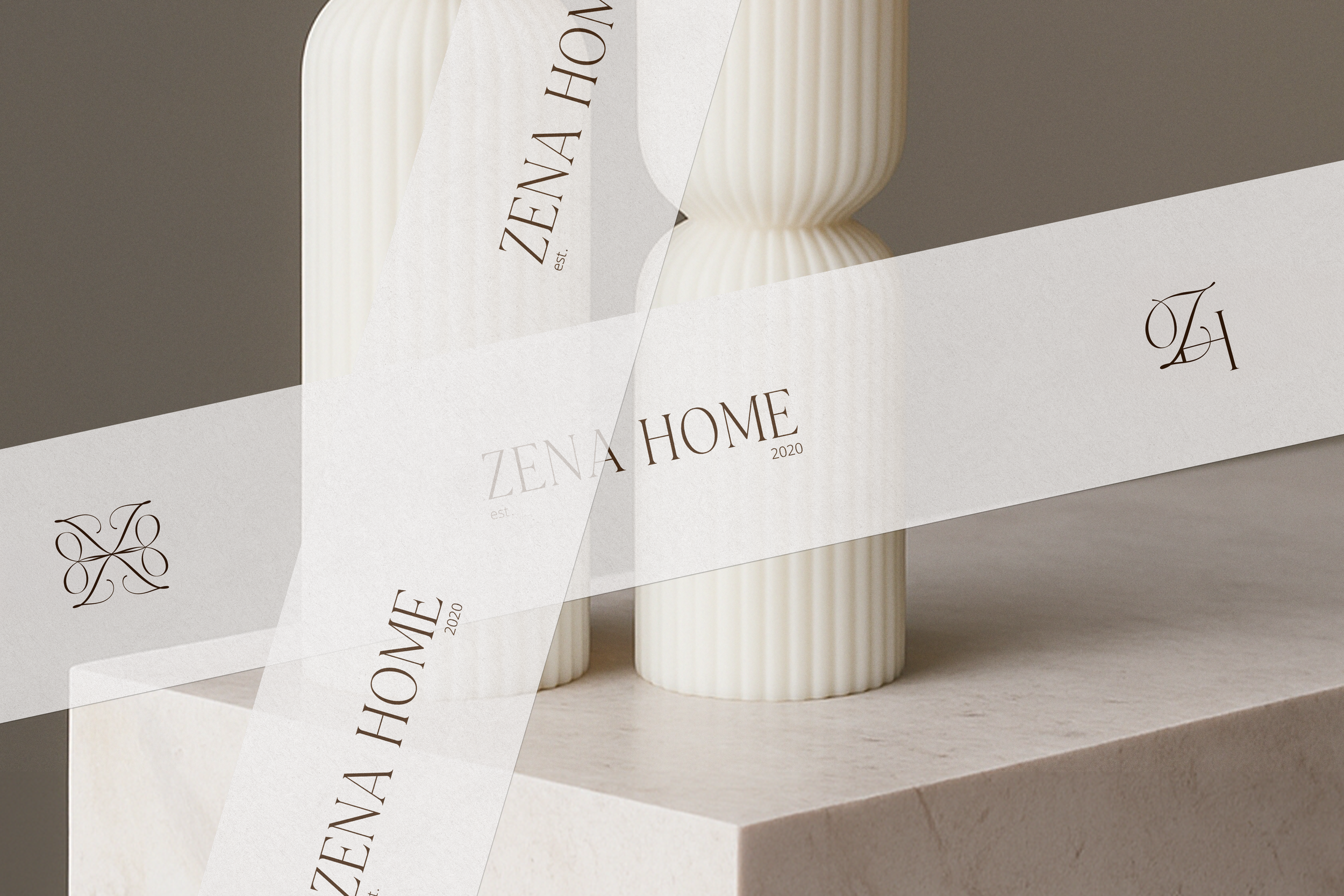 A white ribbon with the Zena Home logo design displayed and decorative secondary logos, wrapped around two handmade ribbed candles, displayed on a light-colored stone surface.
