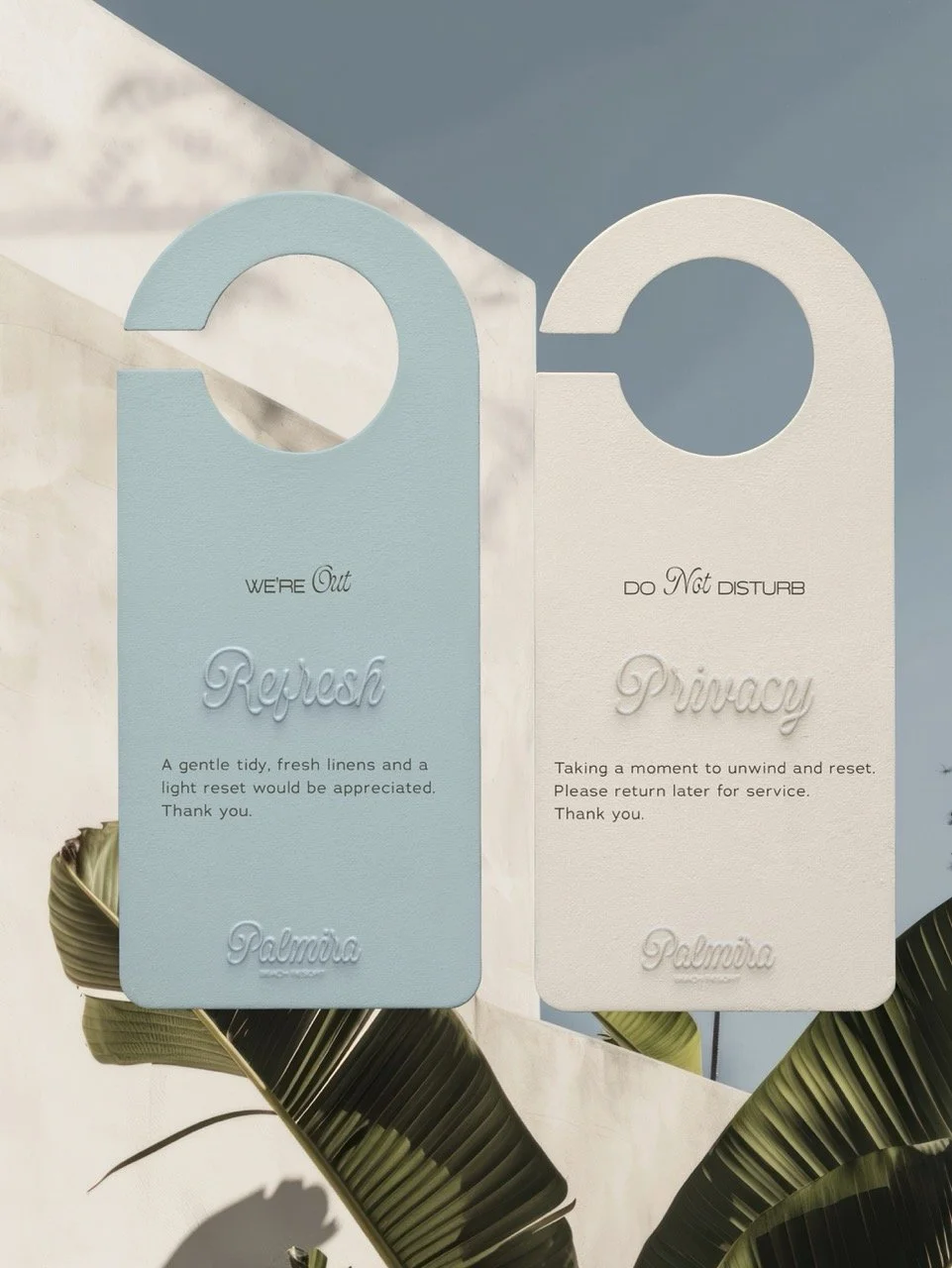 Set of two door hangers, one blue labeled 'We're Out Refast' and one white labeled 'Do Not Disturb Privacy,' hanging on a hook with green tropical leaves nearby.