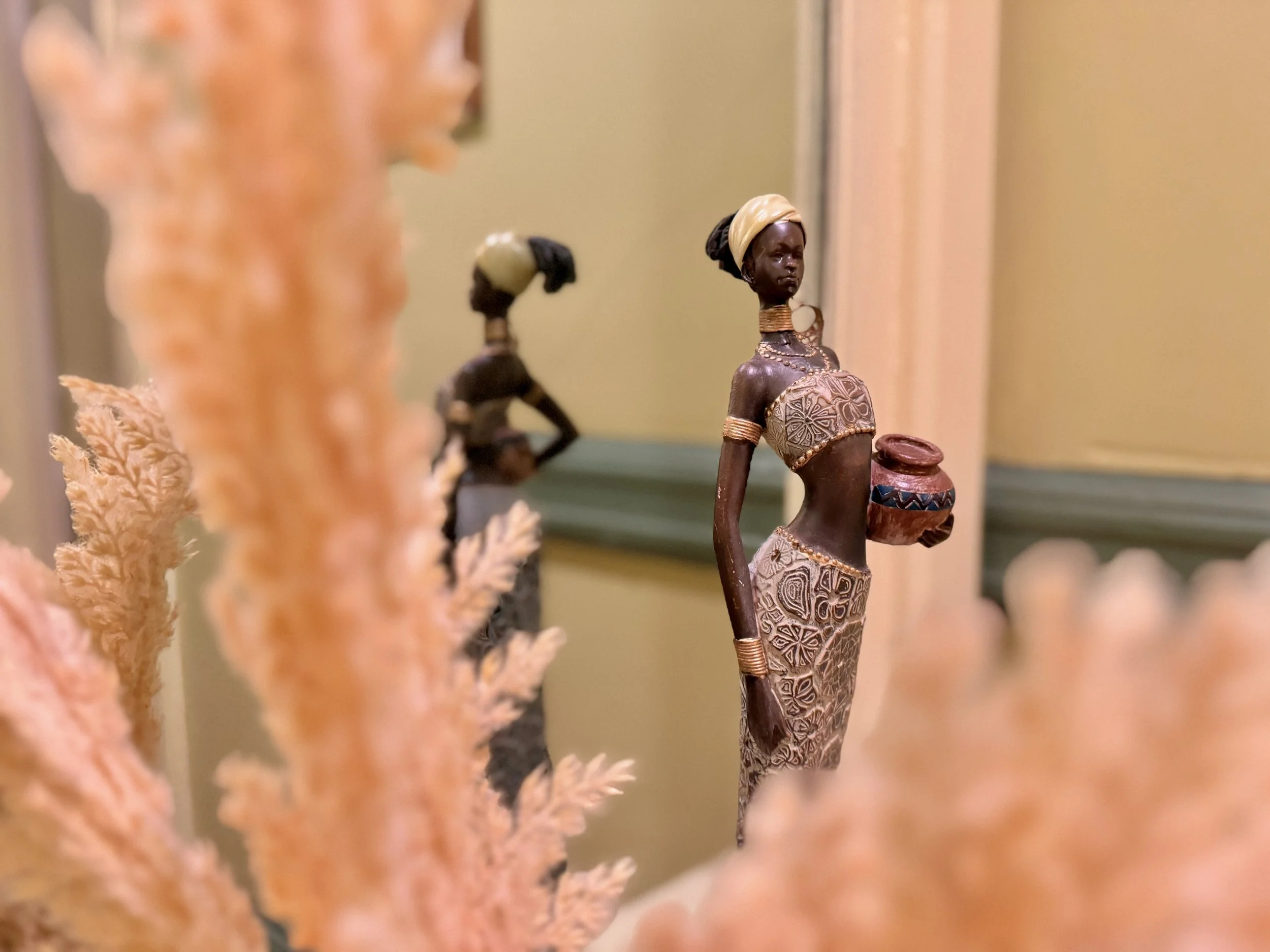 Decorative figurine of an African woman holding a bowl, dressed in patterned clothing and jewelry, with dried plants in the foreground.