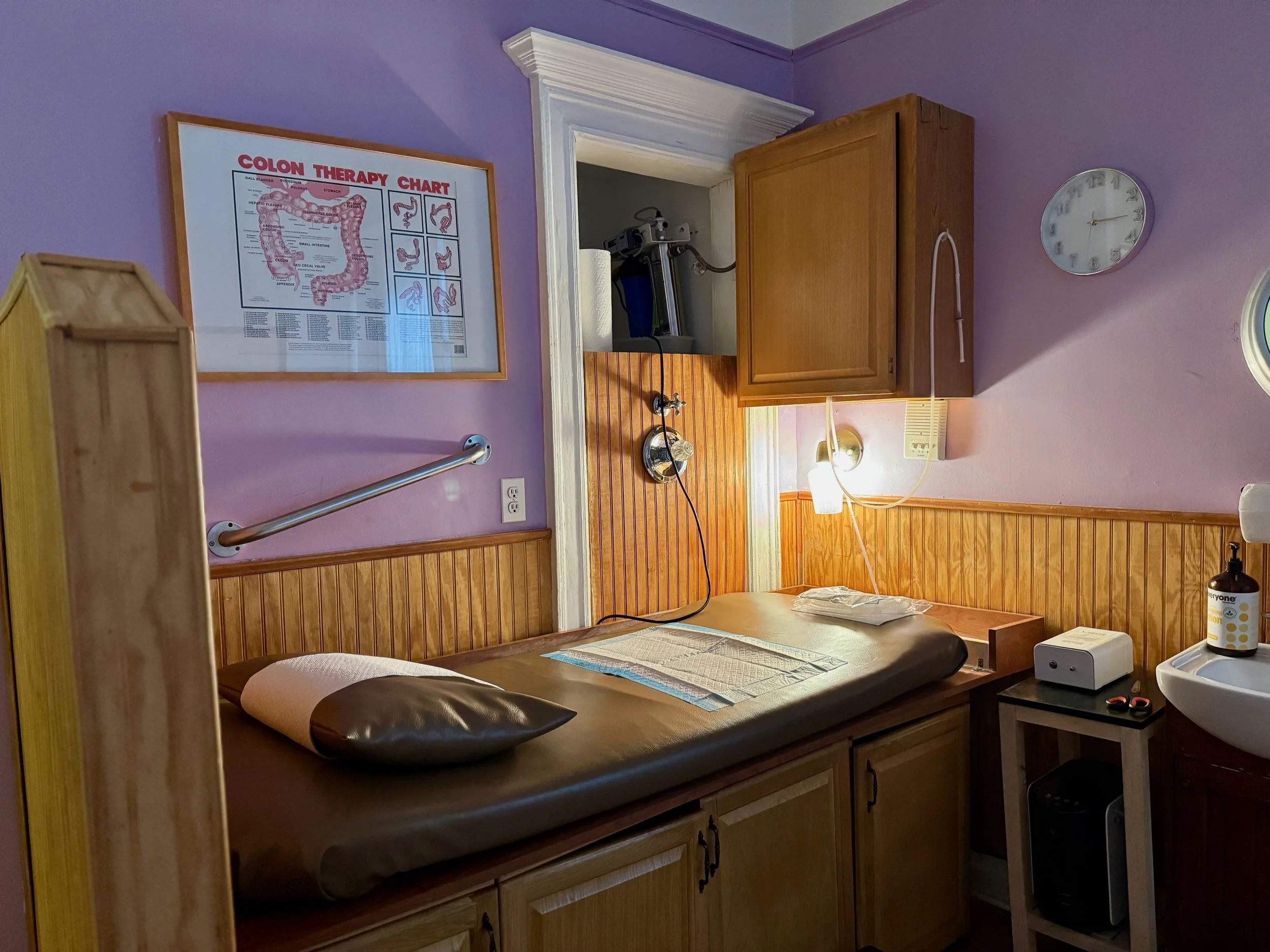 A medical examination room with purple walls, a wooden cabinet, a clock, and a colon therapy chart on the wall. There is an examination bed with a pillow and a sheet, a small table with medical supplies, and a hand sanitizer bottle.