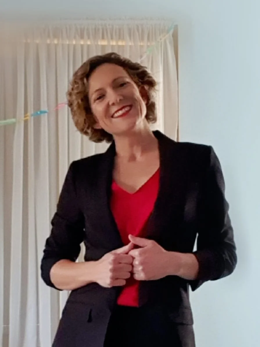 Photo of Ruth Conwell, she is a woman with curly blonde/brown hair, wearing a black blazer over a red top, smiling and standing in front of white curtains.