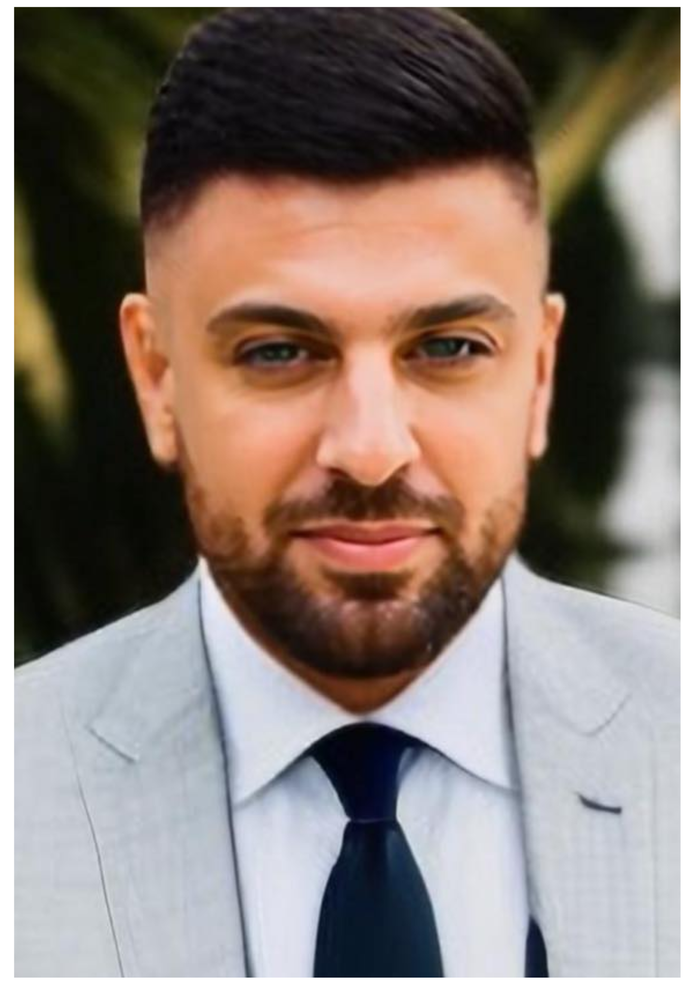 Close-up of Andy Kotzoulas, he is a man in a gray suit and dark tie with short dark hair, beard, and moustache, outdoors with blurred greenery background.