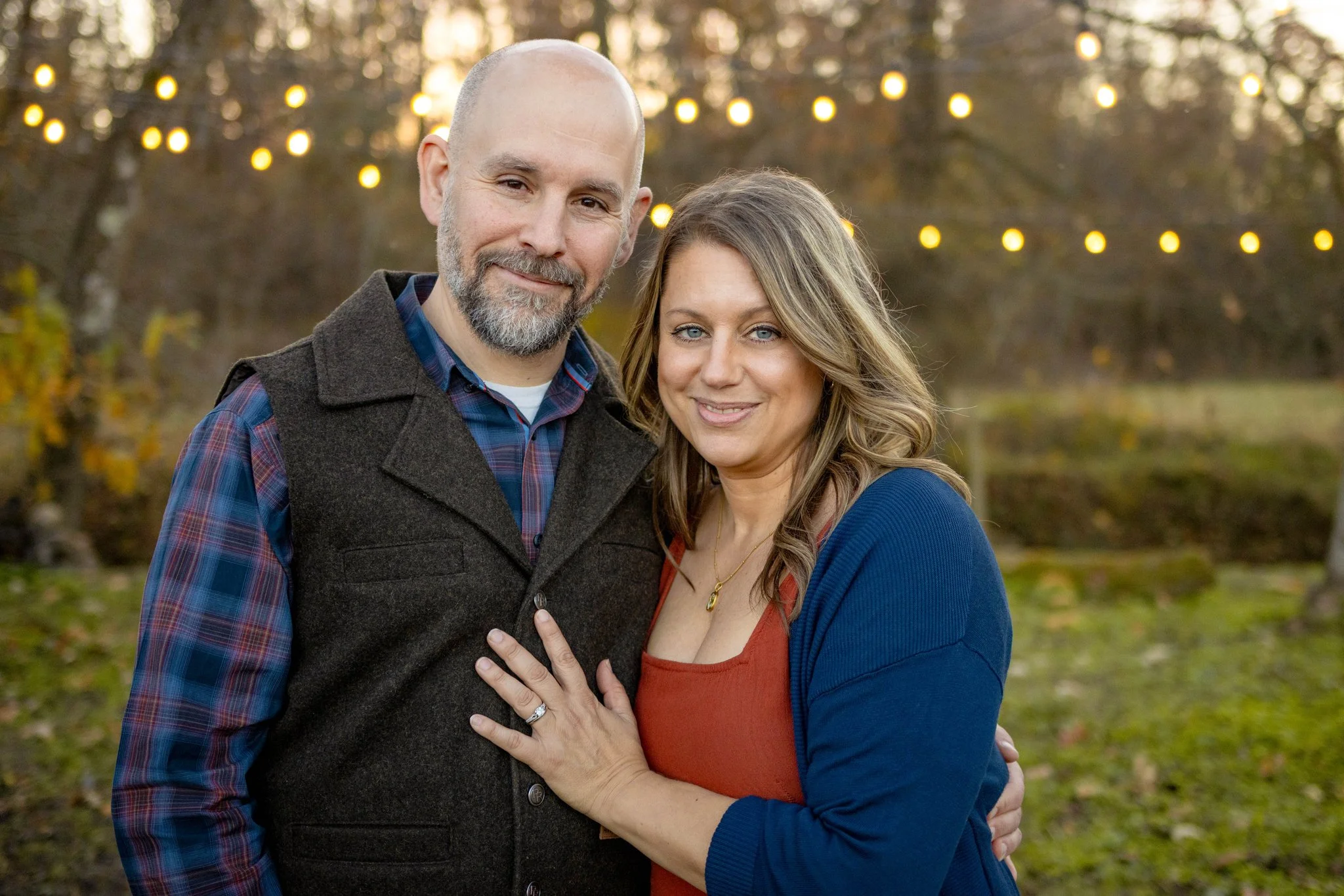 A happy couple stands outdoors with string lights overhead, autumn trees in the background, and the woman showing her engagement ring.