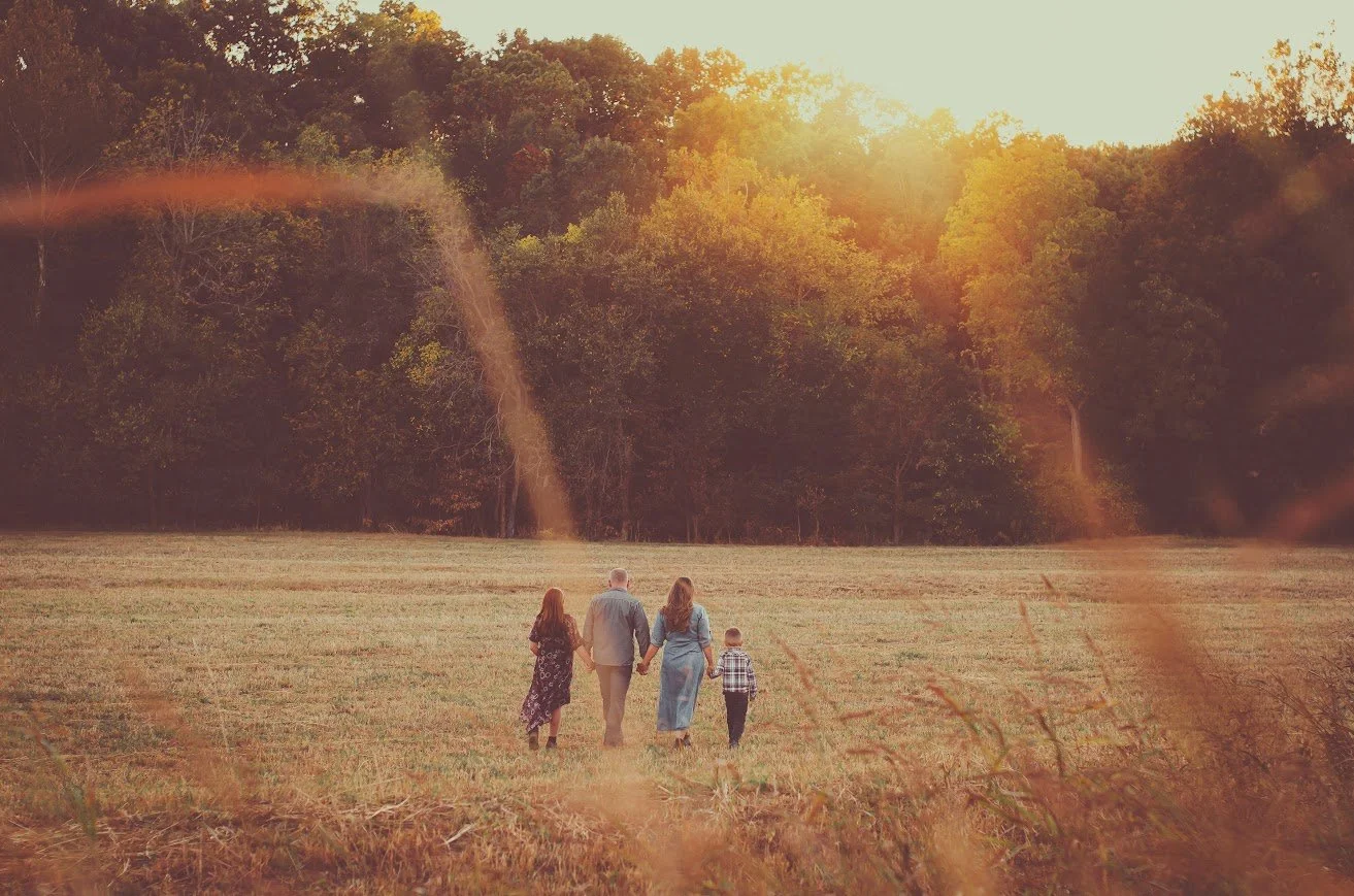 A family of four walking together in a field during sunset, with trees in the background and sunlight filtering through.