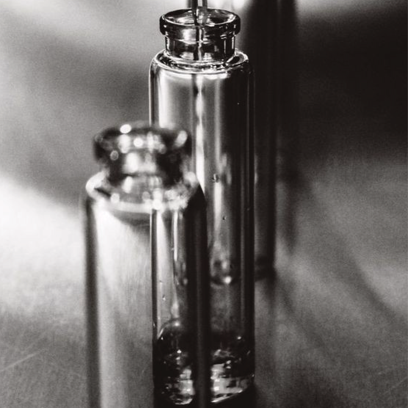 Black and white close-up photo of two glass vials with metal caps, one slightly taller than the other, on a dark surface.