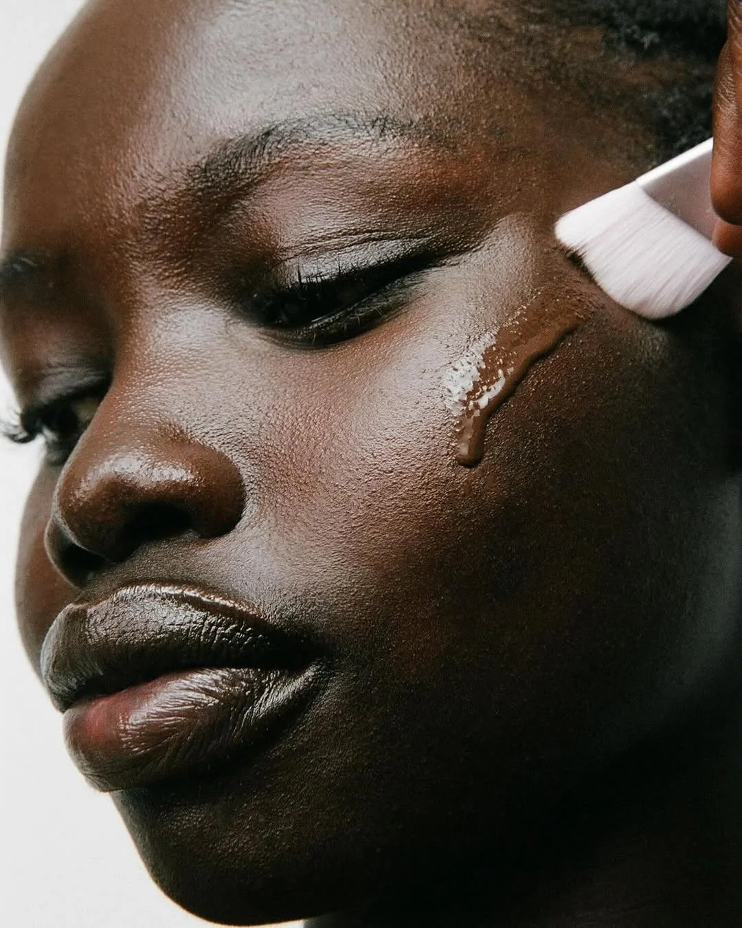A close-up of a woman getting a facial treatment with a white brush and a liquid product on her cheek.