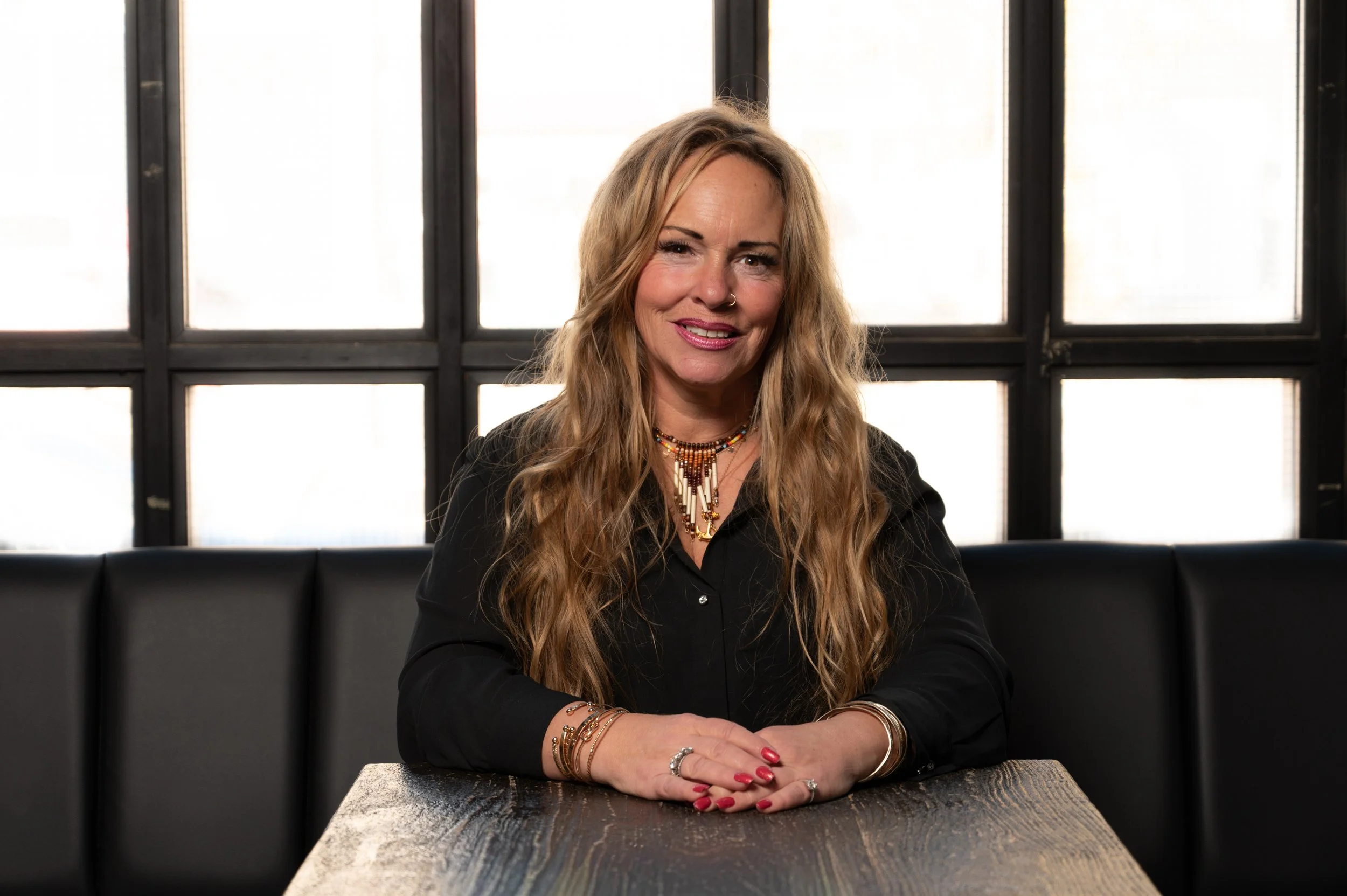 A woman with long wavy blonde hair, wearing a black top and colorful jewelry, sitting at a wooden table in front of a windowed wall with a grid pattern, smiling at the camera.