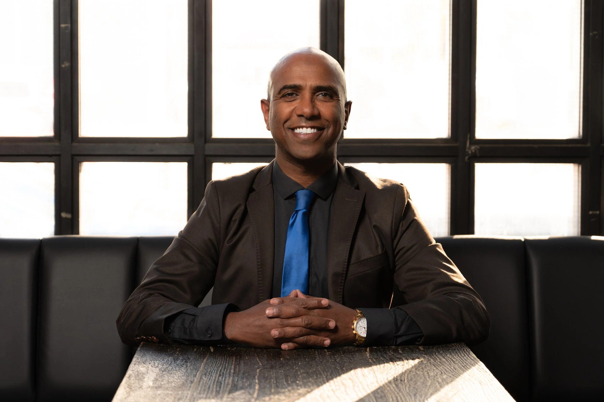 A smiling man in a suit with a blue tie sitting at a table in front of a large window in an office or restaurant.