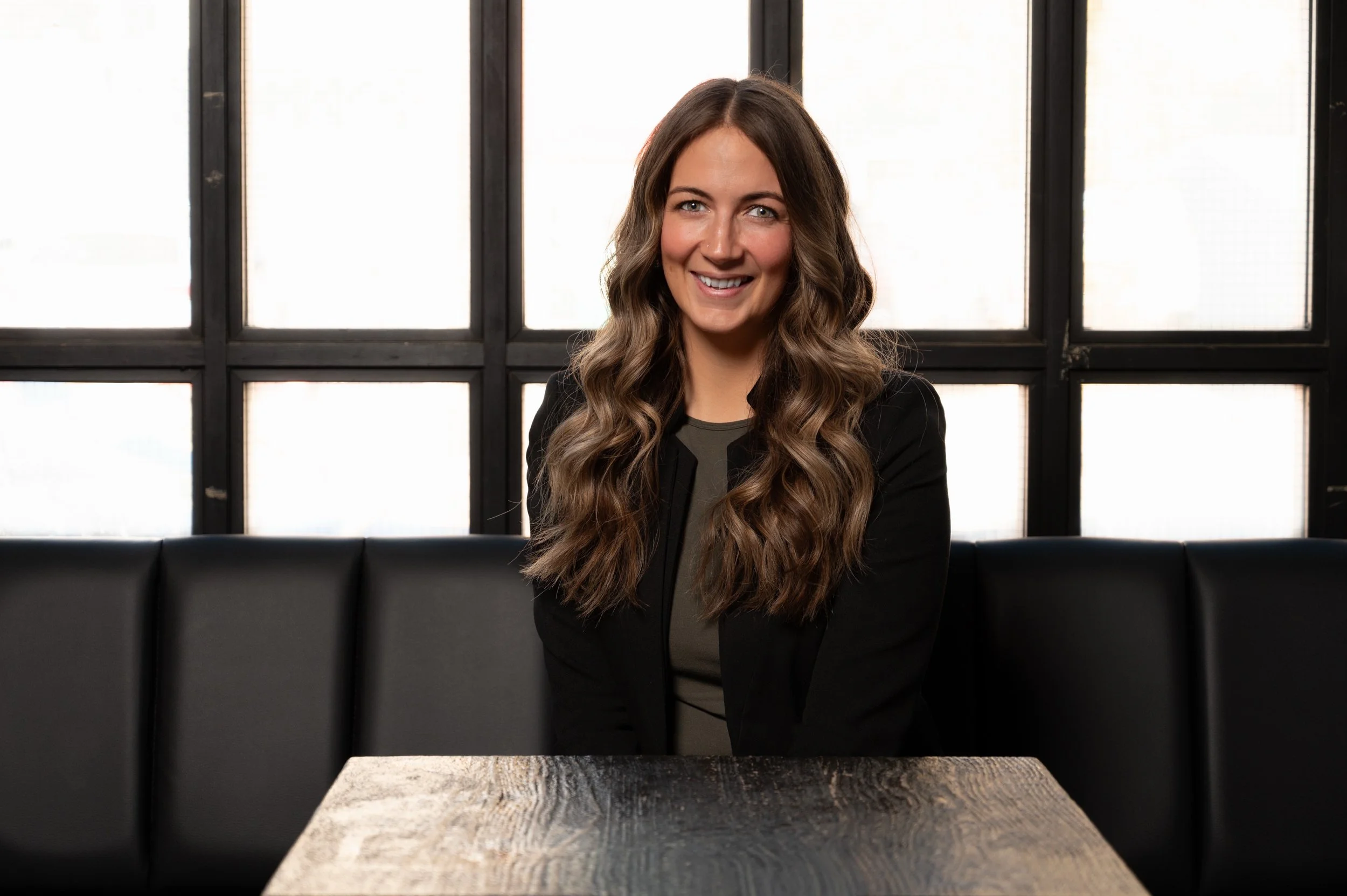 A woman with long, wavy brown hair, smiling, sitting in a black booth at a wooden table in front of a window with multiple panes.