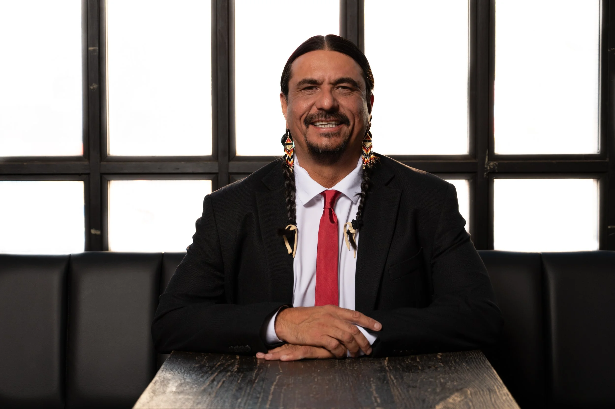 A man with long braided hair and colorful earrings smiling, sitting at a wooden table in front of a windowed background.