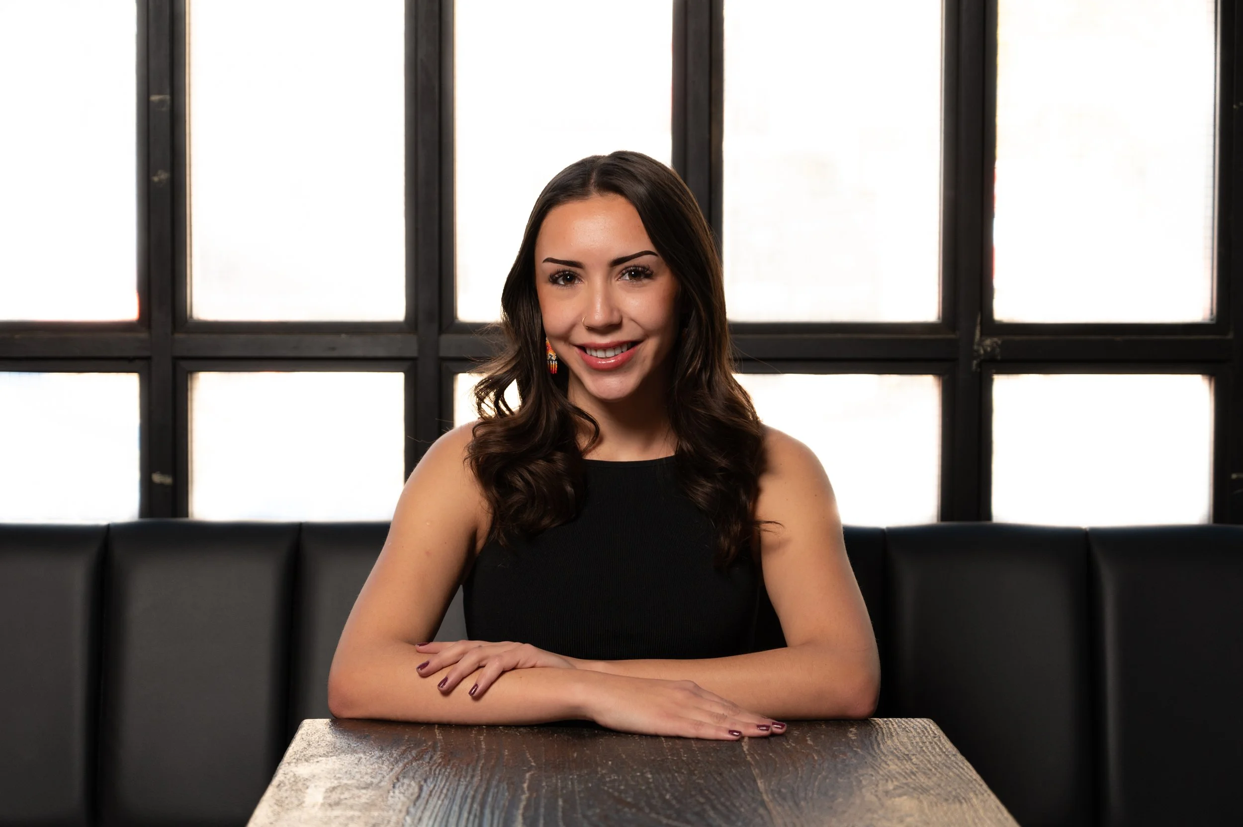 A young woman with long wavy dark hair, wearing a black sleeveless top, seated at a wooden table in a room with large black-framed windows in the background.