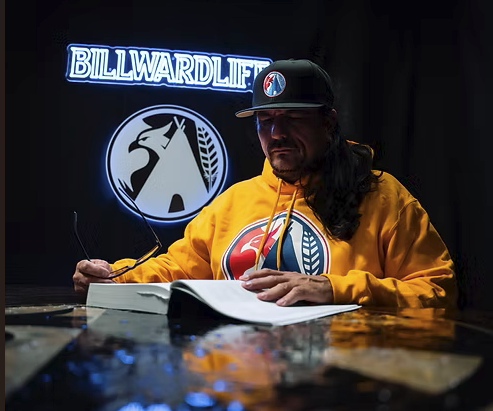 A man with long hair and a beard wears a black cap and a yellow hoodie with a logo. He is sitting at a table with a book or magazine and manning glasses. Behind him are illuminated signs, one says 'BILLWARDLIFE' and another with a graphic of a practitioner.