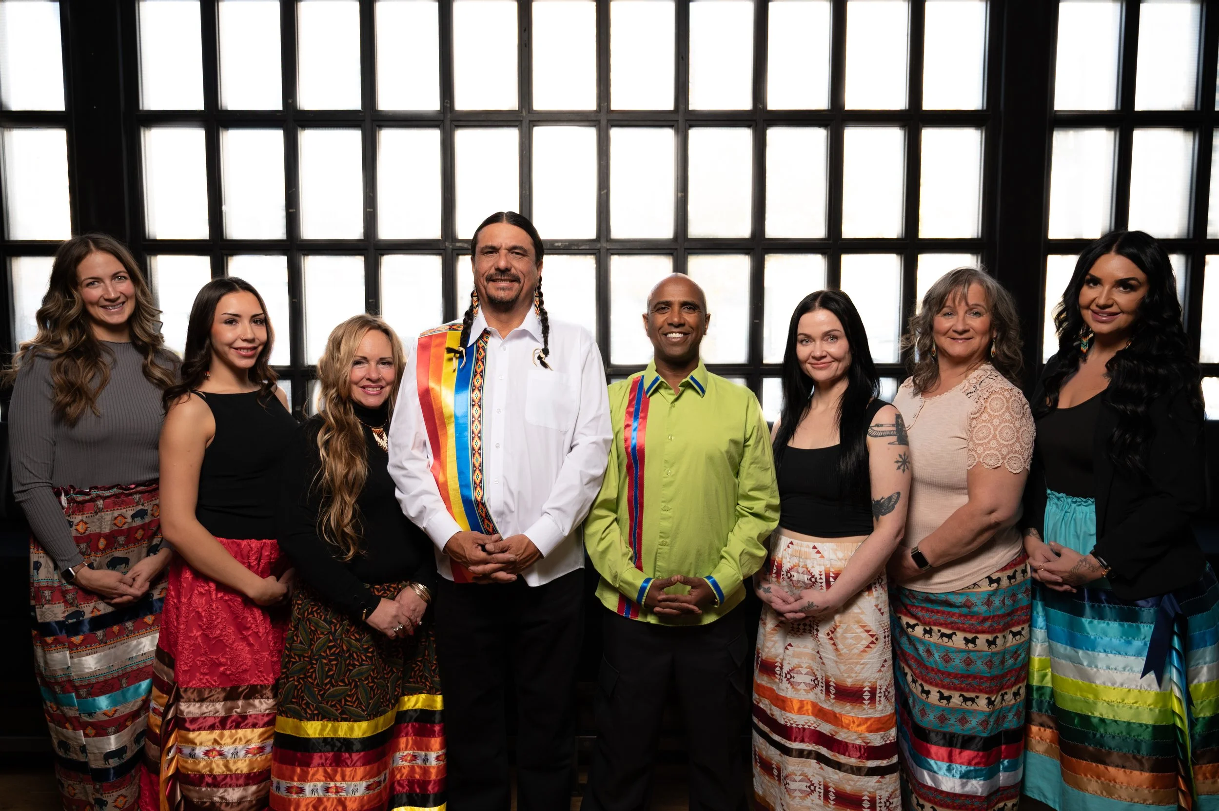 A group of eight diverse people standing in front of a large window, smiling, wearing colorful clothing with traditional patterns.