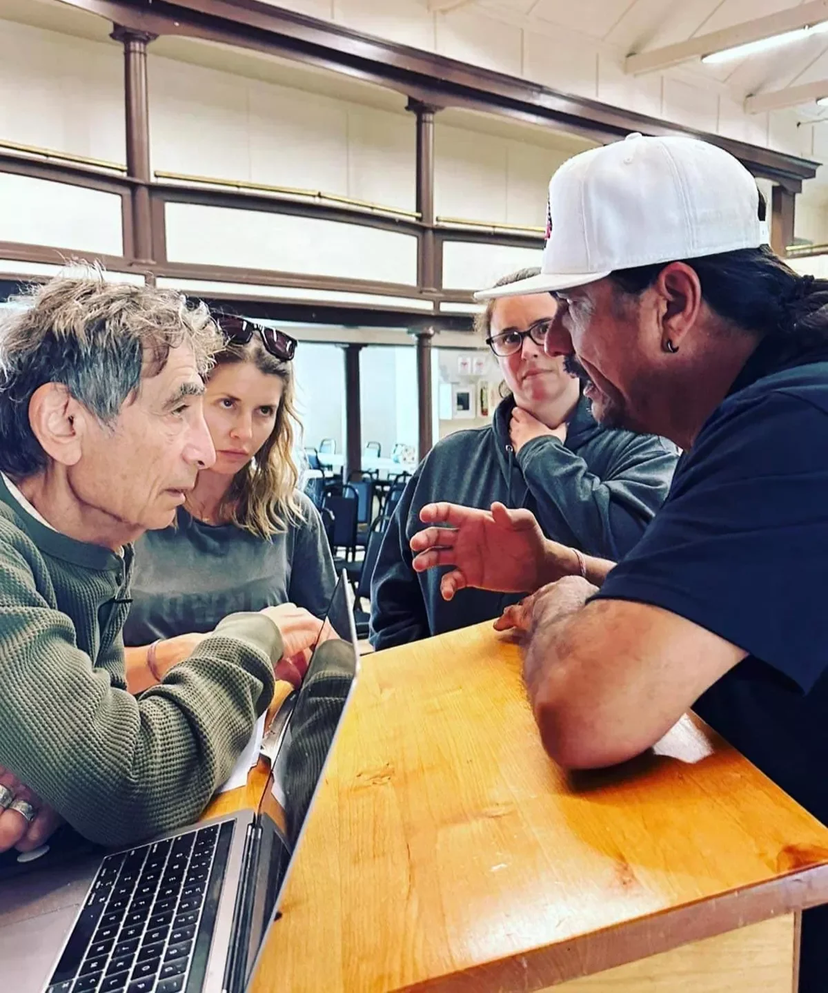 Four people engaged in a serious discussion at a wooden table, with a man in a white cap explaining something to three people, one Gabor Mate the other has glasses and is touching her chin.