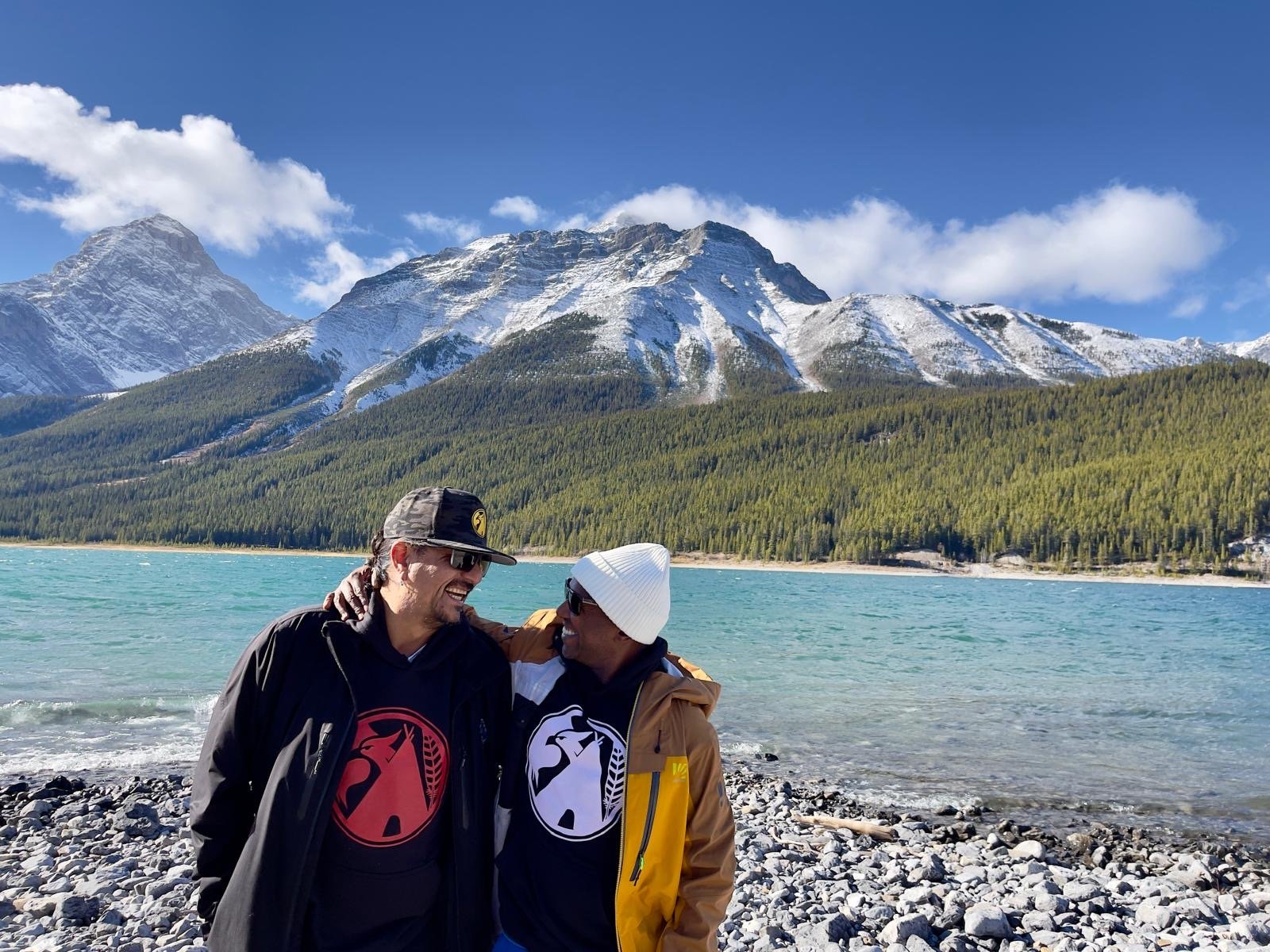 Two men stand close together on a rocky beach by a mountain lake, smiling and embracing each other, with snow-capped mountains and a clear blue sky in the background.
