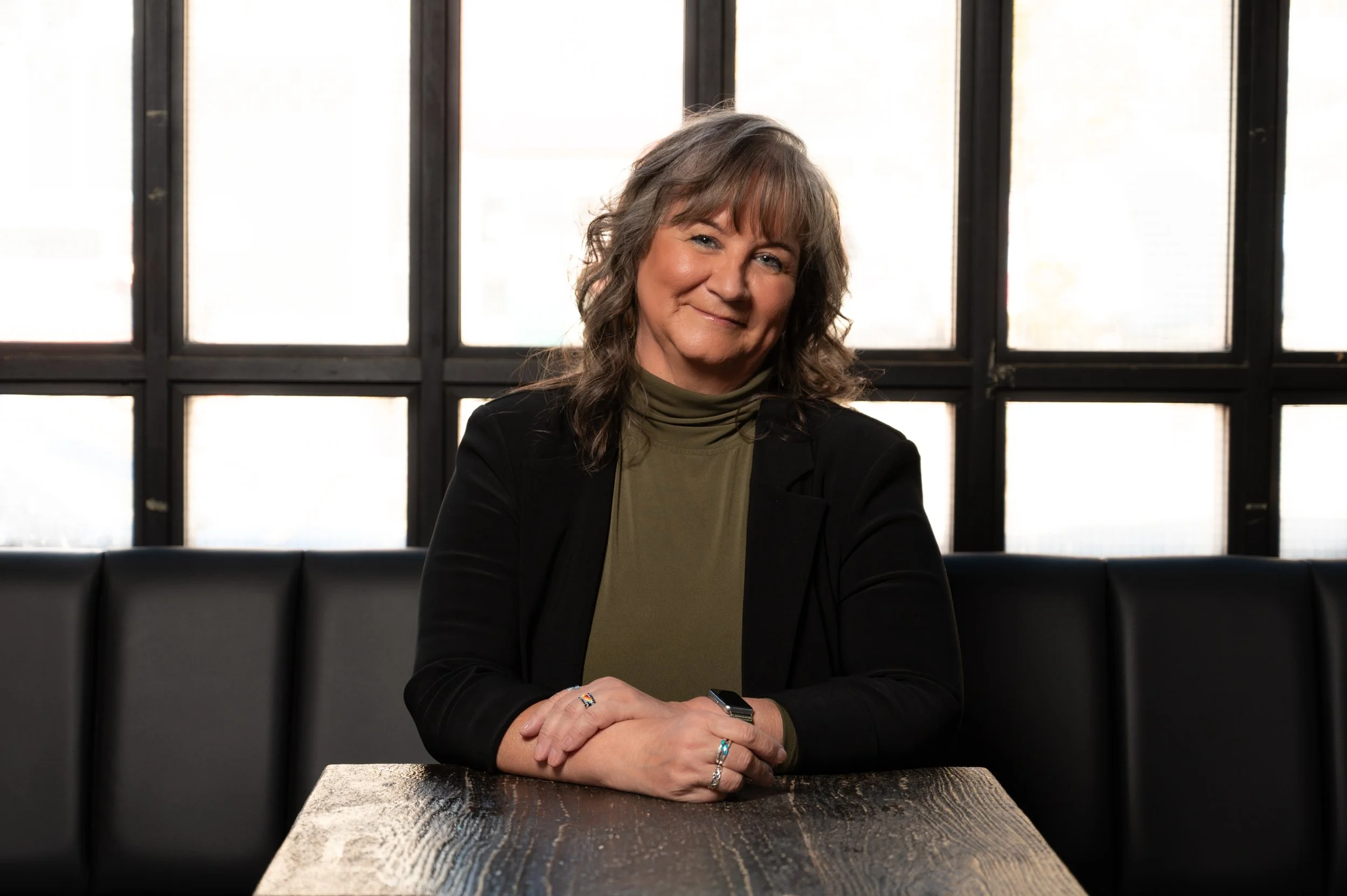 A woman with shoulder-length wavy hair, wearing a black blazer and olive green turtleneck, sitting at a wooden table in a modern room with large window panes behind her.
