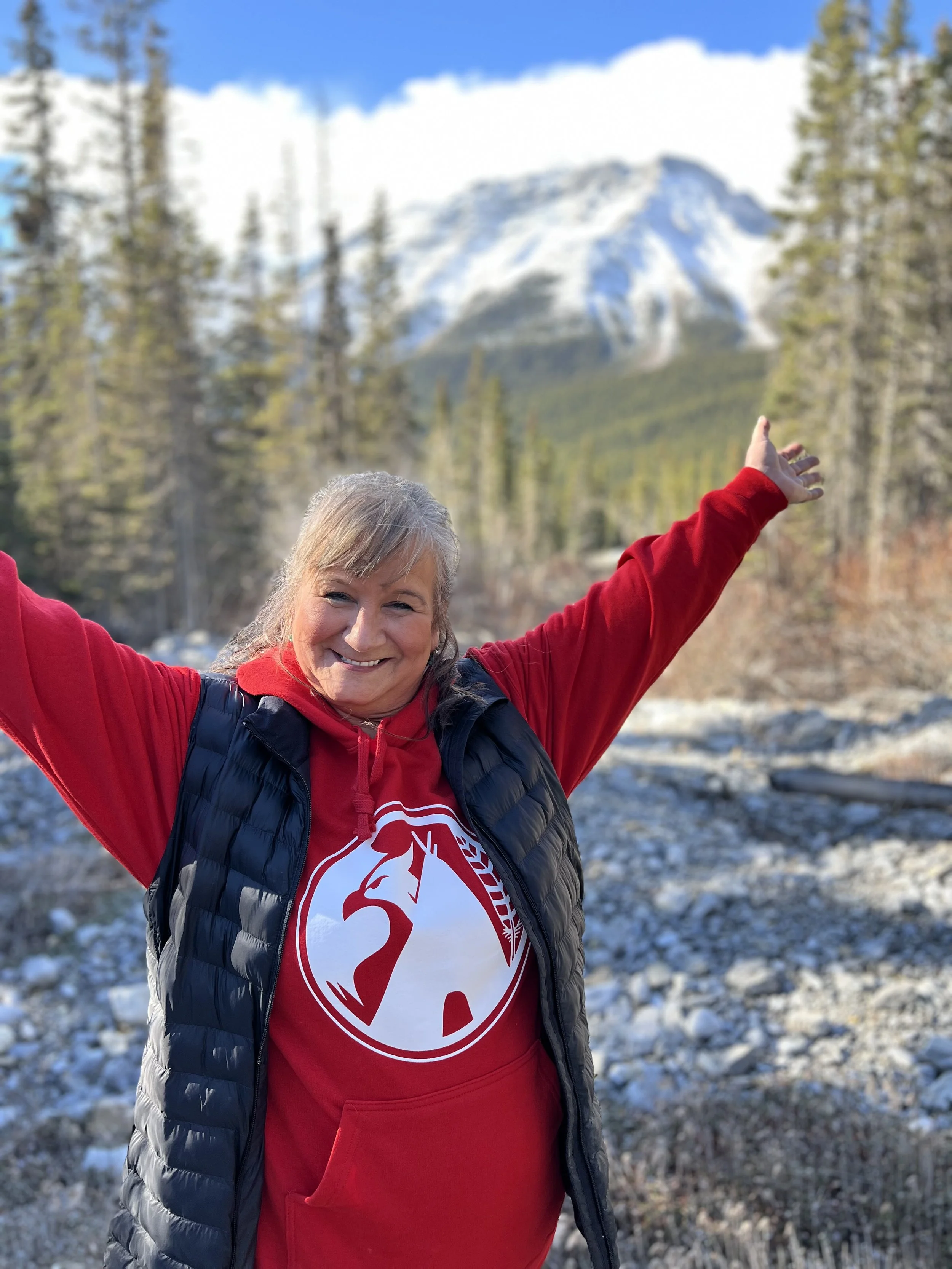A smiling woman in a red sweatshirt and black puffer vest standing outdoors with arms raised, with a snowy mountain and trees in the background.