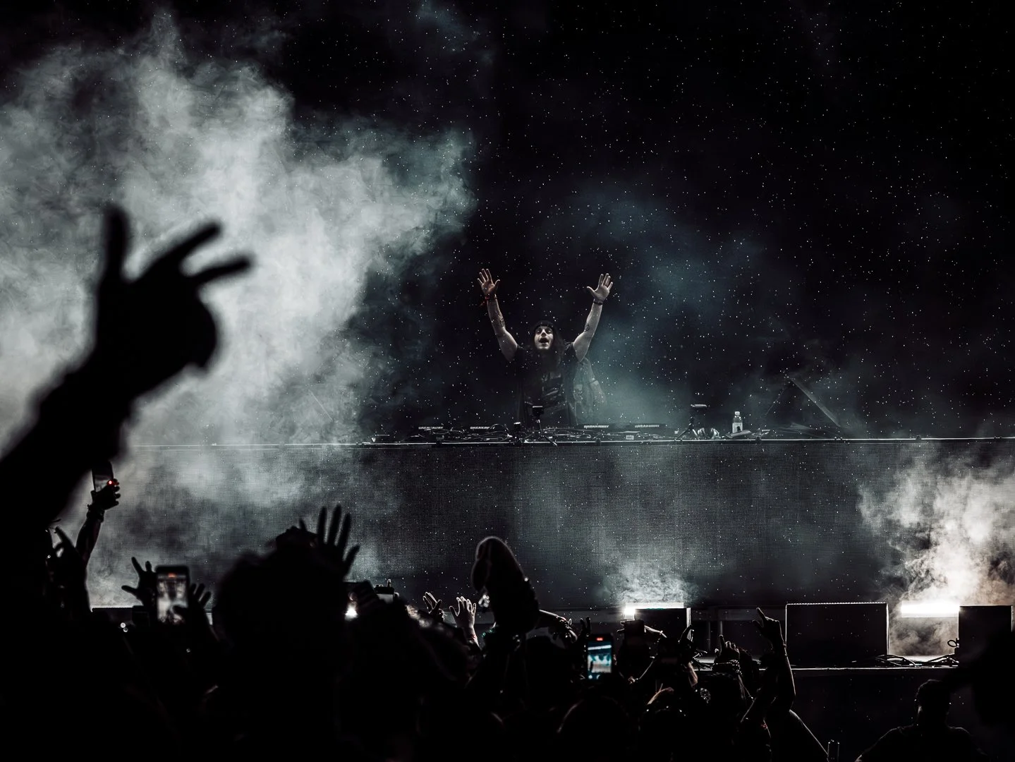 DJ performing on stage with raised hands, smoke effects, and starry background, crowd cheering with hands and phones in the foreground.