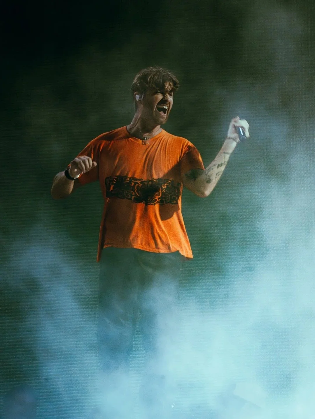 A man singing and dancing on stage, wearing an orange t-shirt and holding a microphone, with smoke and stage lighting in the background.