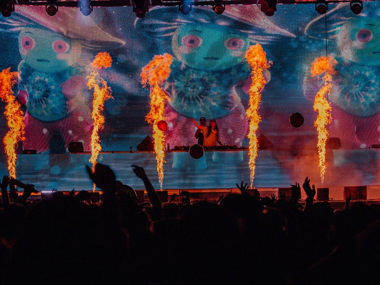 A DJ performs on stage with flames shooting up from the floor, a colorful digital backdrop of animated faces, and an audience with raised hands.