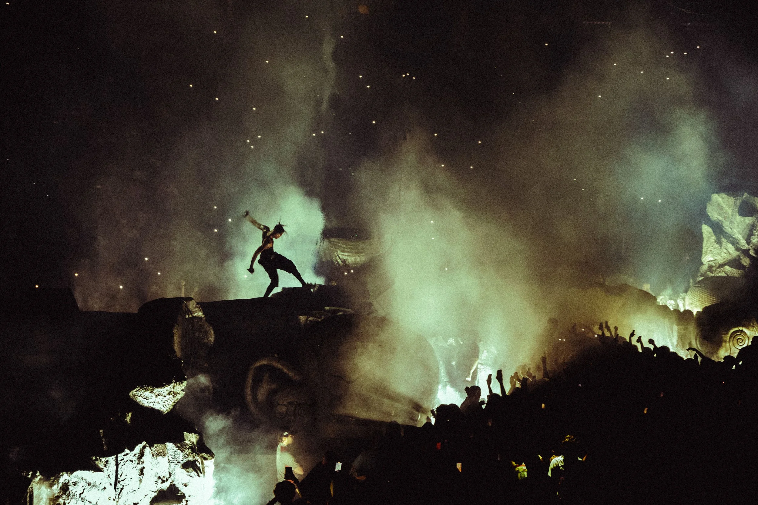 Person performing a stunt or dance atop a large, surreal sculpture of a face at night, with a crowd watching and smoke or fog in the air, illuminated by green lights.