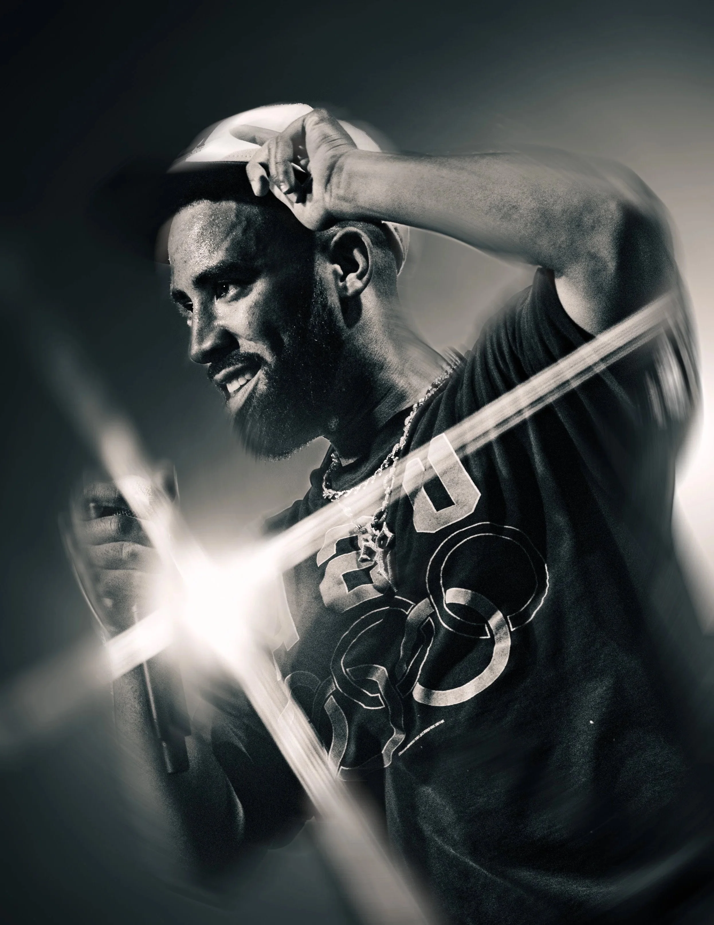 A black and white photo of a young man with a beard, smiling and putting on a cap, with motion blur effects.