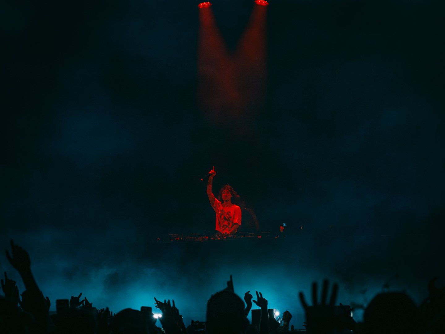 A DJ performing at a concert with red lighting, raising one hand, crowd with hands raised and taking photos below.