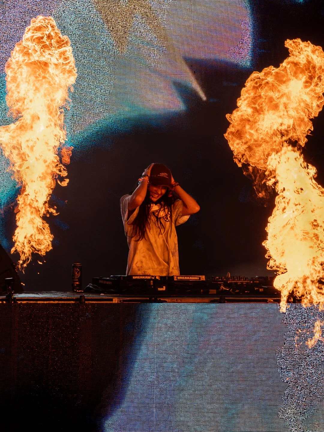 A DJ performing on stage surrounded by flames with a colorful LED screen in the background.