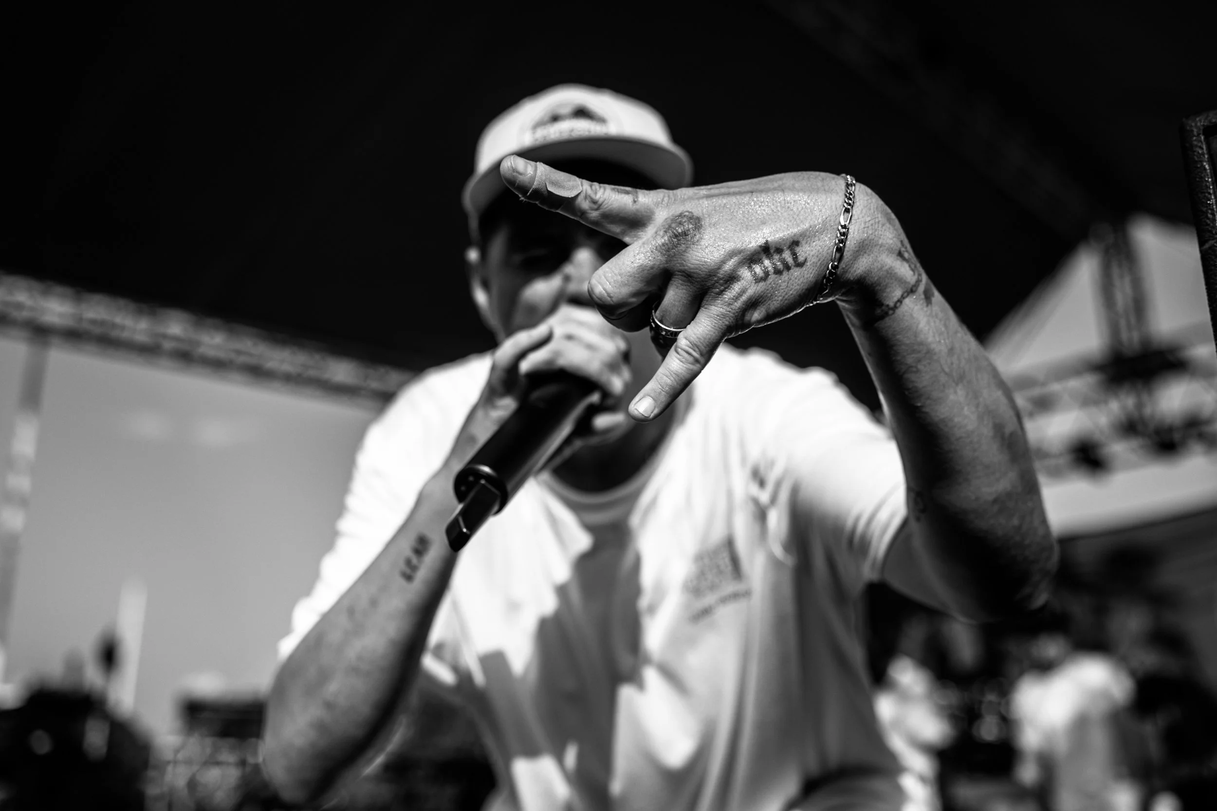 A person wearing a white shirt and cap, holding a microphone and making a hand gesture with their middle finger extended, during an outdoor event.