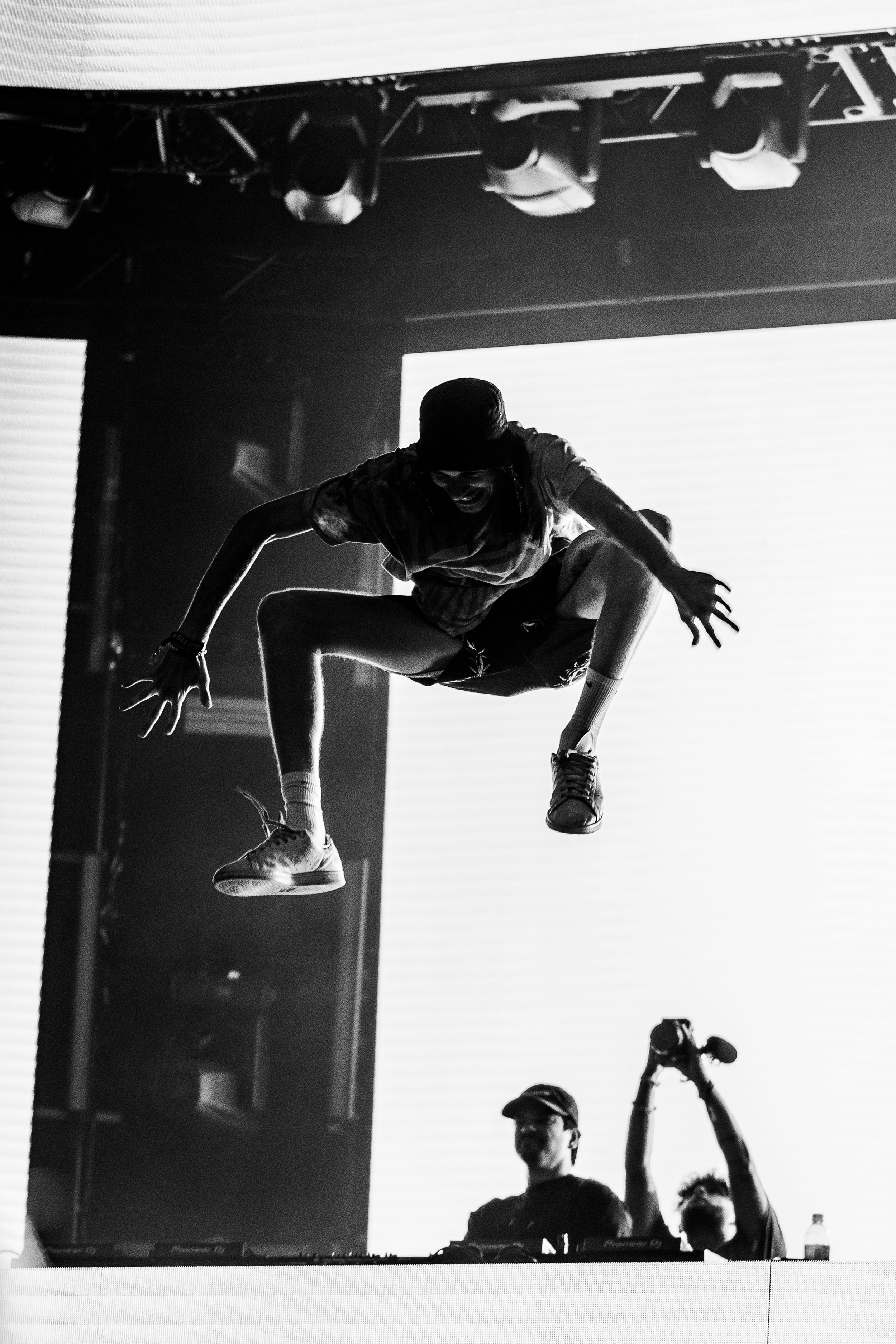 A person jumps high in the air during a performance with stage lights and a large screen in the background, while a DJ stands at a turntable below.