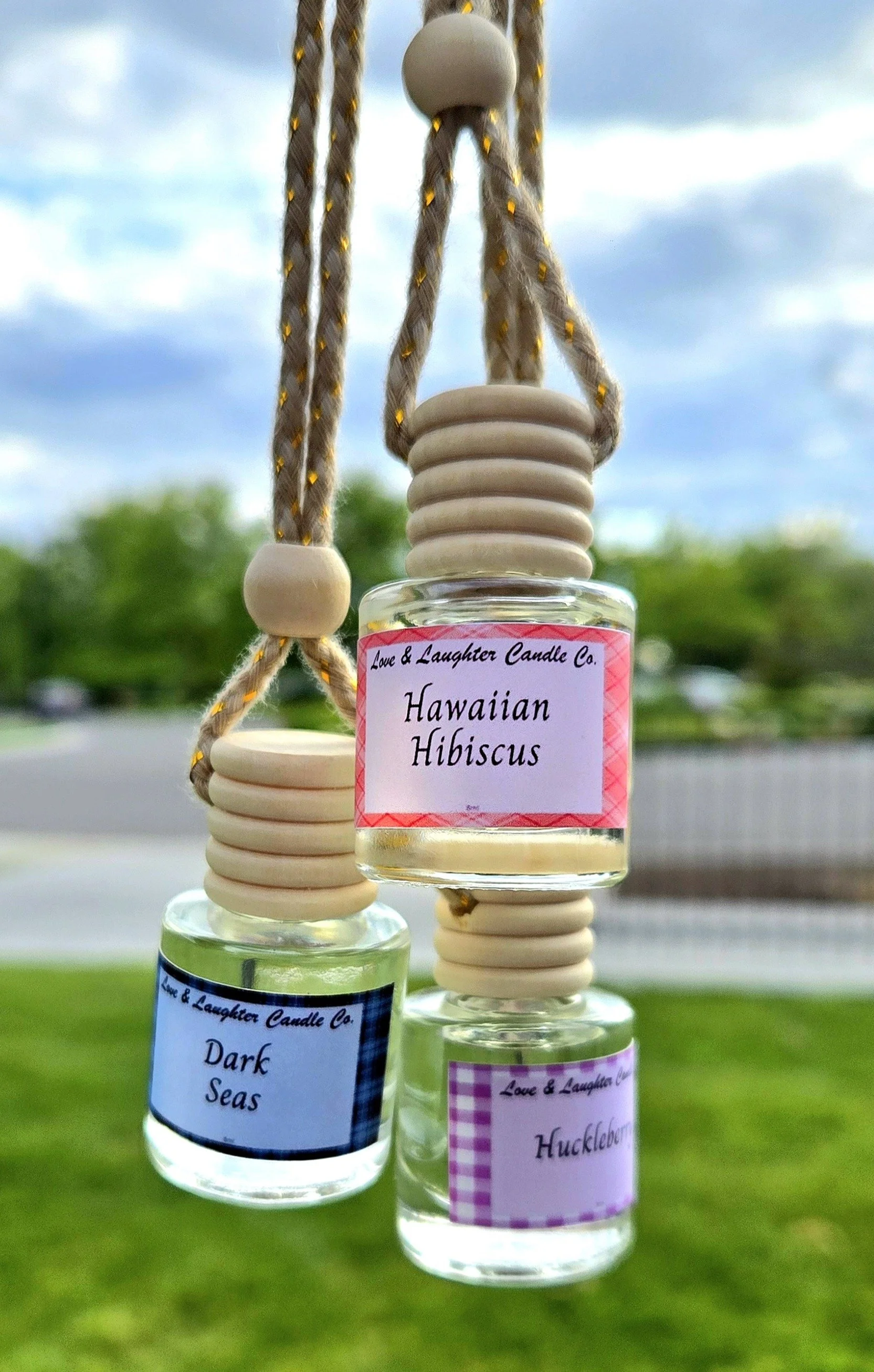 Three small glass candles with wooden lids and lavender-colored labels hanging from a beige and yellow twisted cord outdoors, with trees and a cloudy sky in the background.
