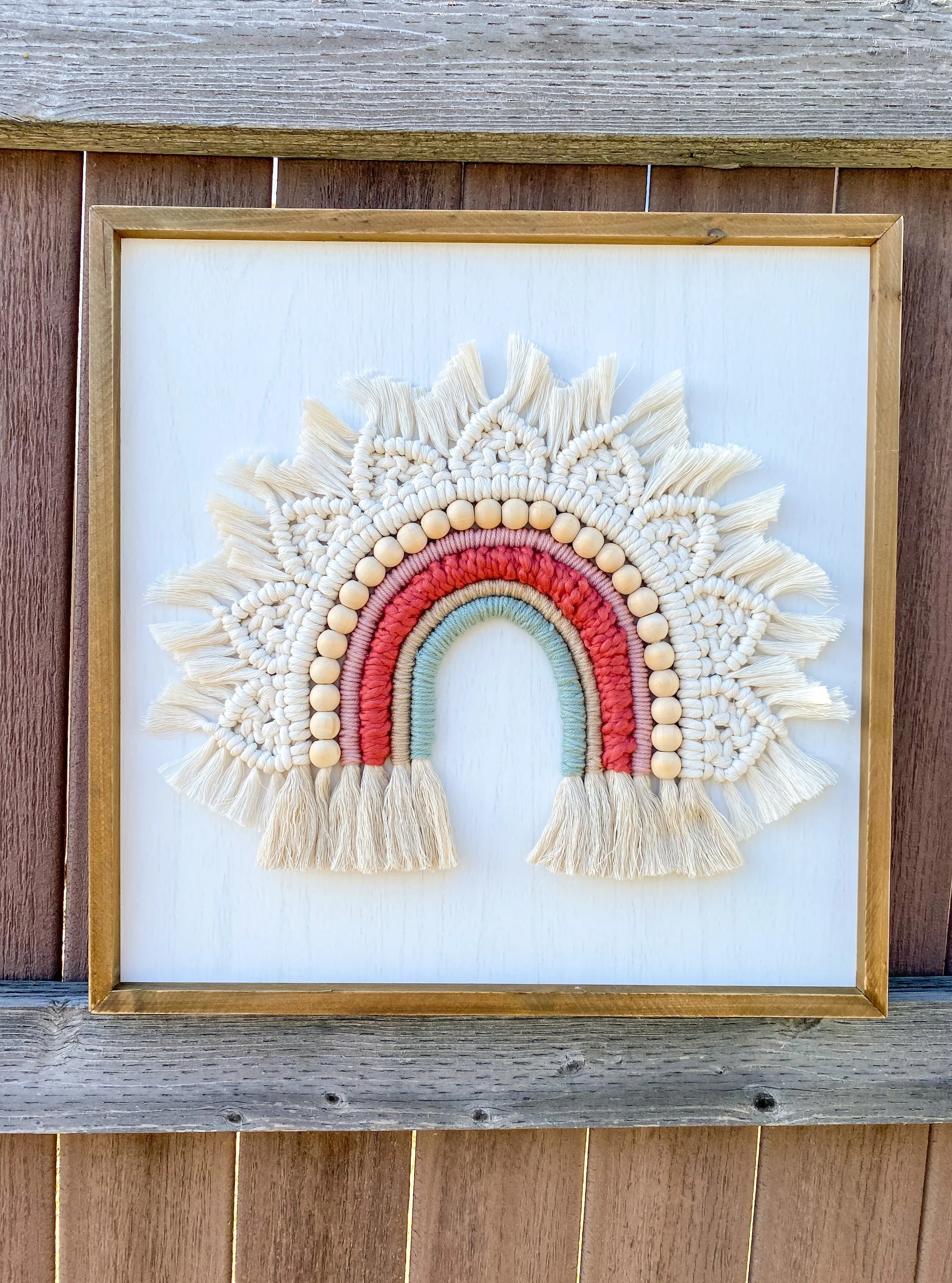Decorative wall hanging of a rainbow with pink, green, and red yarn surrounded by beige tassels and beads, framed in wood.