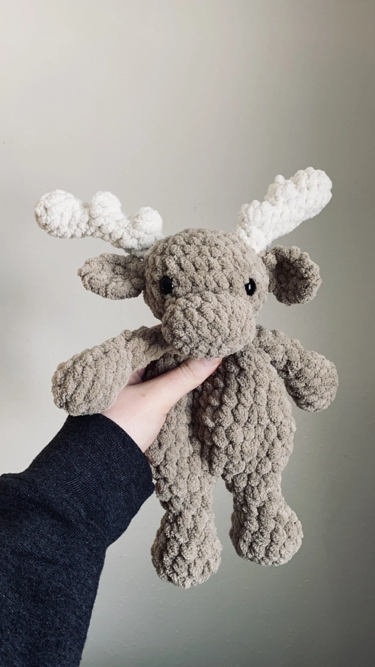 A plush reindeer toy with a soft, bumpy texture, a large nose, black eyes, and white antlers being held against a plain wall.
