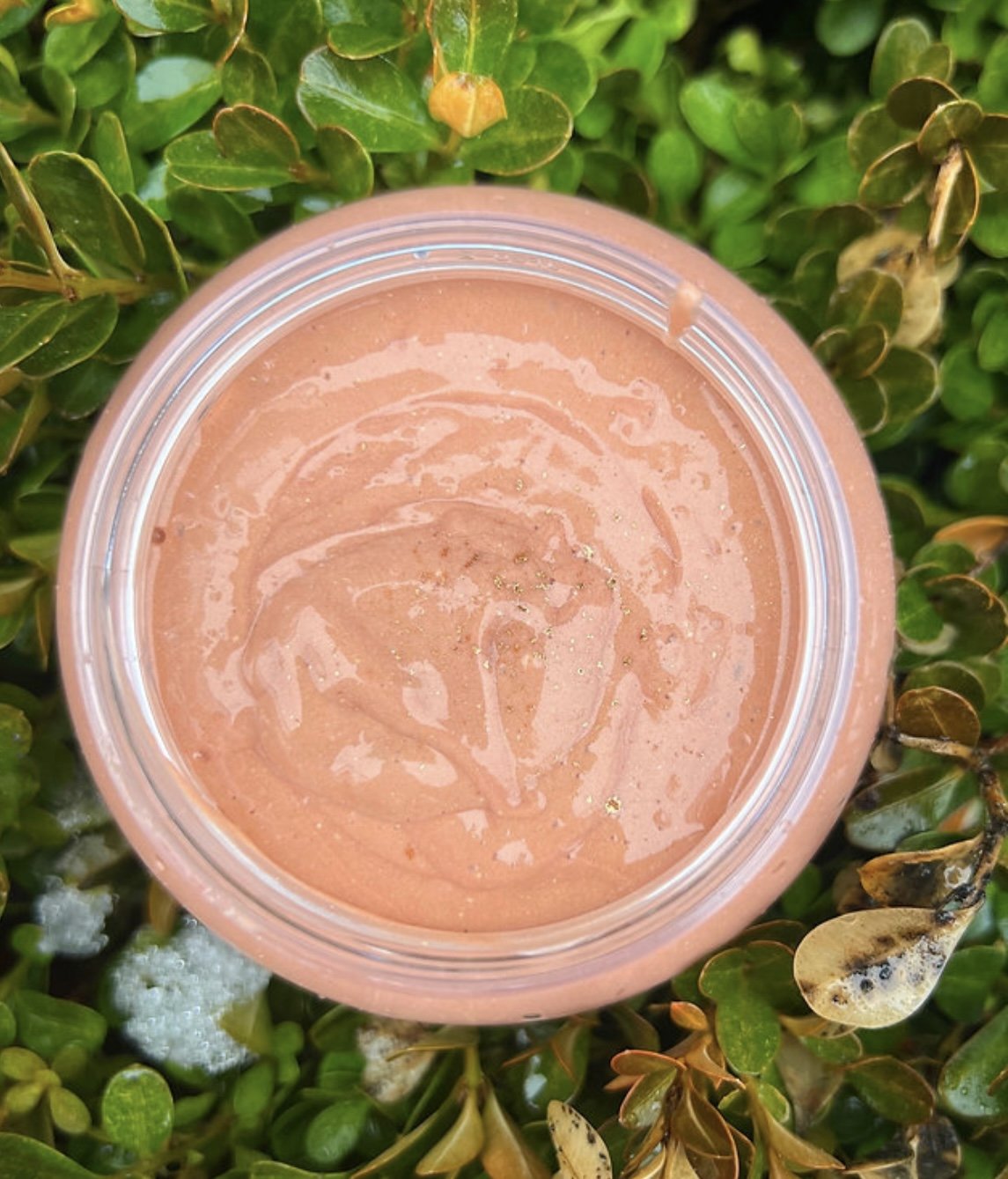 Open jar of peach-colored body cream or lotion with a swirl pattern on top, surrounded by green leaves.