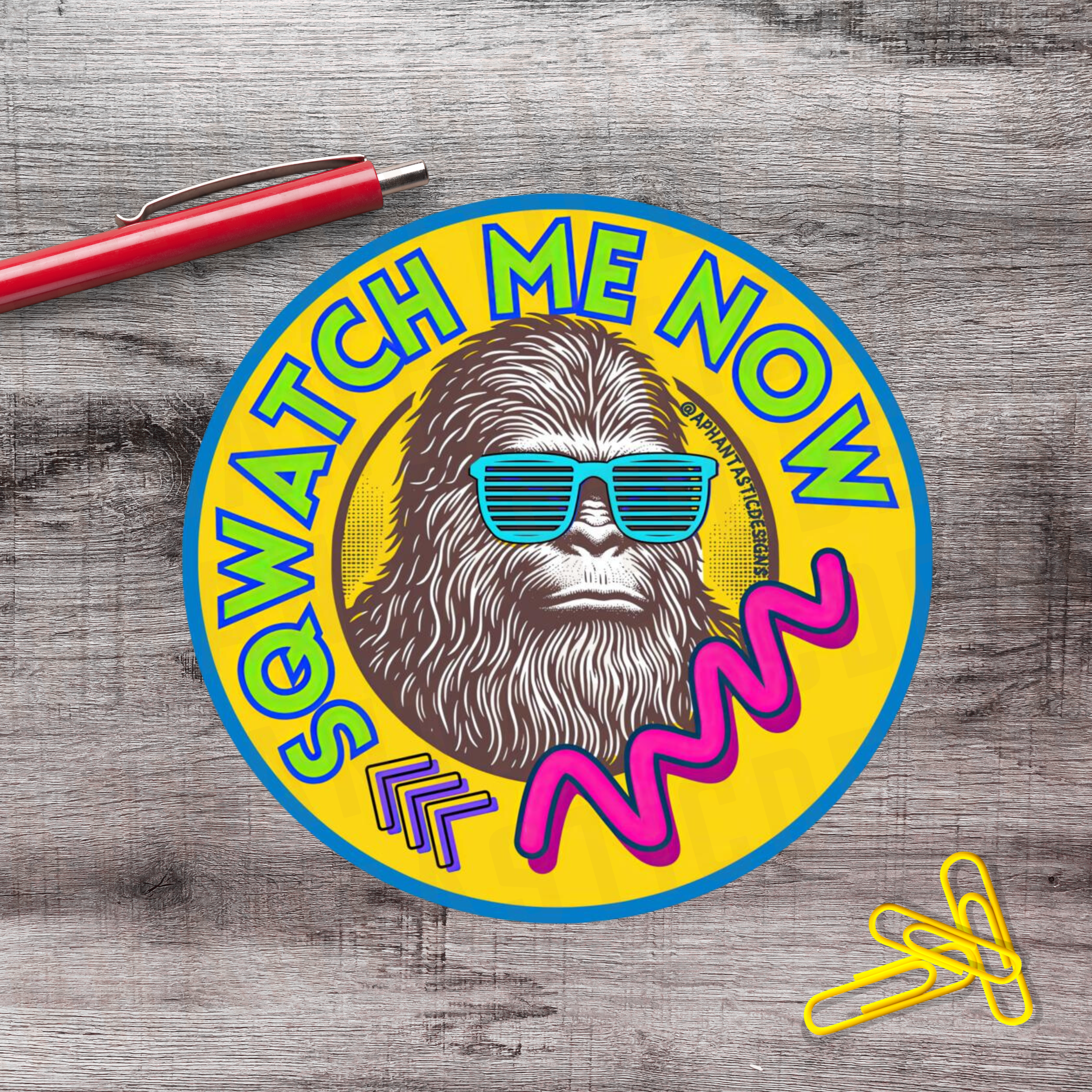 Colorful round sign with a beard man wearing sunglasses and the text 'SO WHY WATCH ME NOW' in bright colors, placed on a wooden surface with a red pen and yellow paperclips nearby.