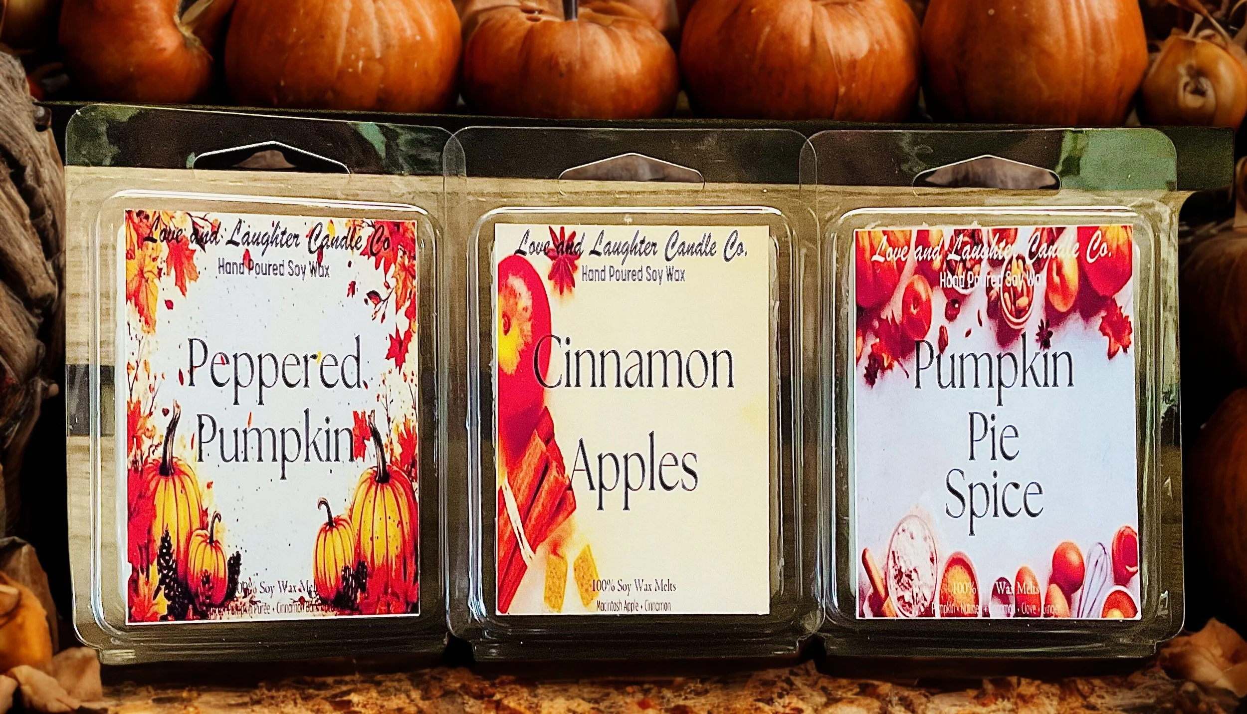 Three wax melt packages labeled 'Peppered Pumpkin,' 'Cinnamon Apples,' and 'Pumpkin Pie Spice,' with autumn-themed designs, placed in front of pumpkins.