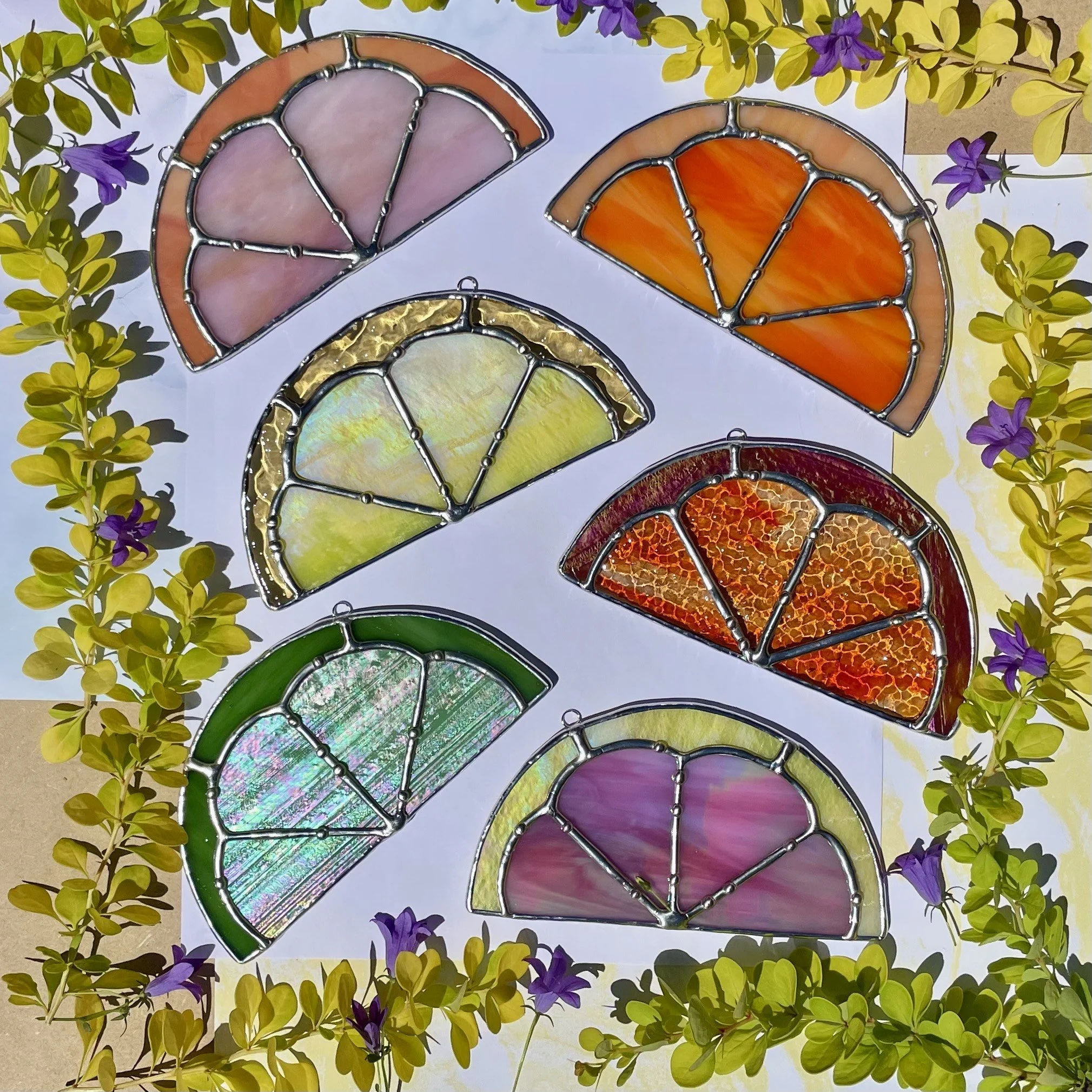 Six stained glass semicircular ornaments arranged on white paper, featuring pink, orange, yellow, green, purple, and multicolored designs, surrounded by greenery and purple flowers.