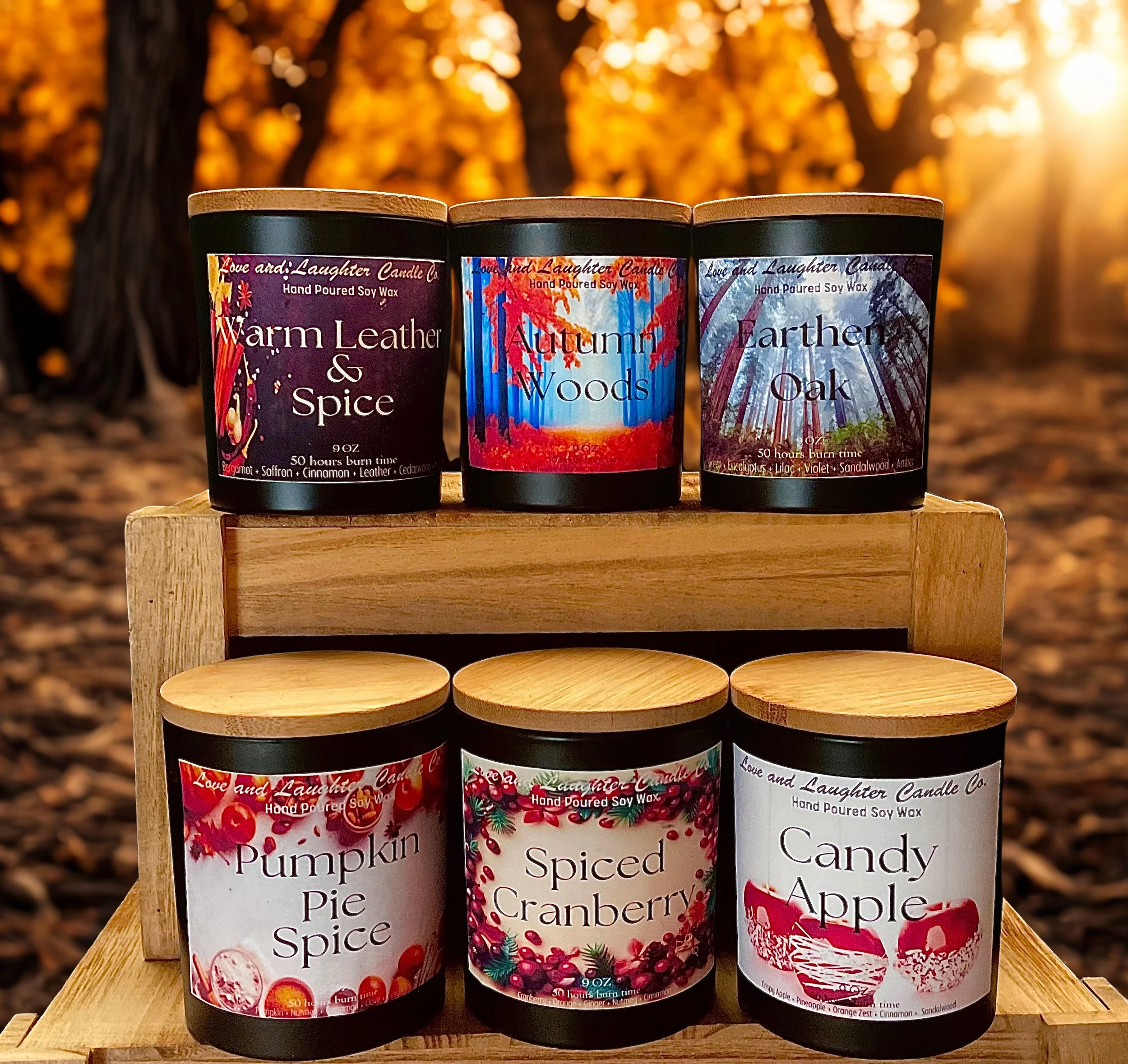 Six candles with decorative labels, arranged on a wooden stand outdoors with a background of autumn trees and warm sunlight. The candles are labeled 'Warm Leather & Spice,' 'Autumn Woods,' 'Earthed Oak,' 'Pumpkin Pie Spice,' 'Spiced Cranberry,' and 'Candy Apple.'