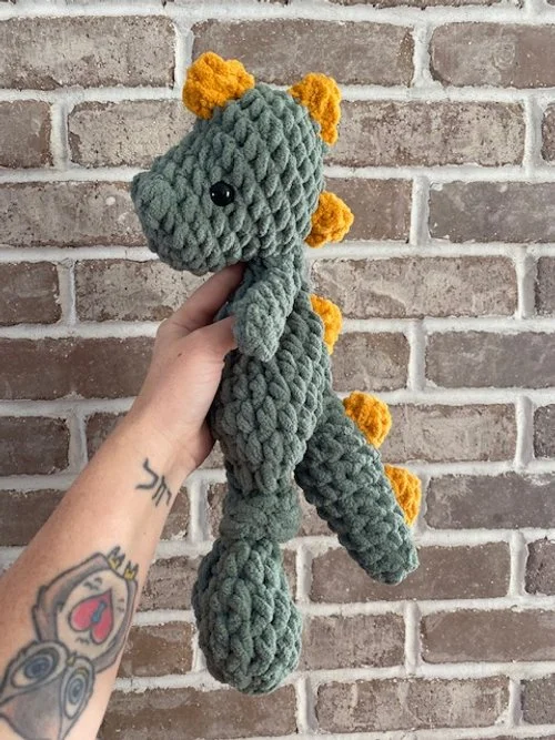 Hand holding a plush, crocheted dinosaur toy with green body, yellow spikes, and a black eye, against a brick wall background.